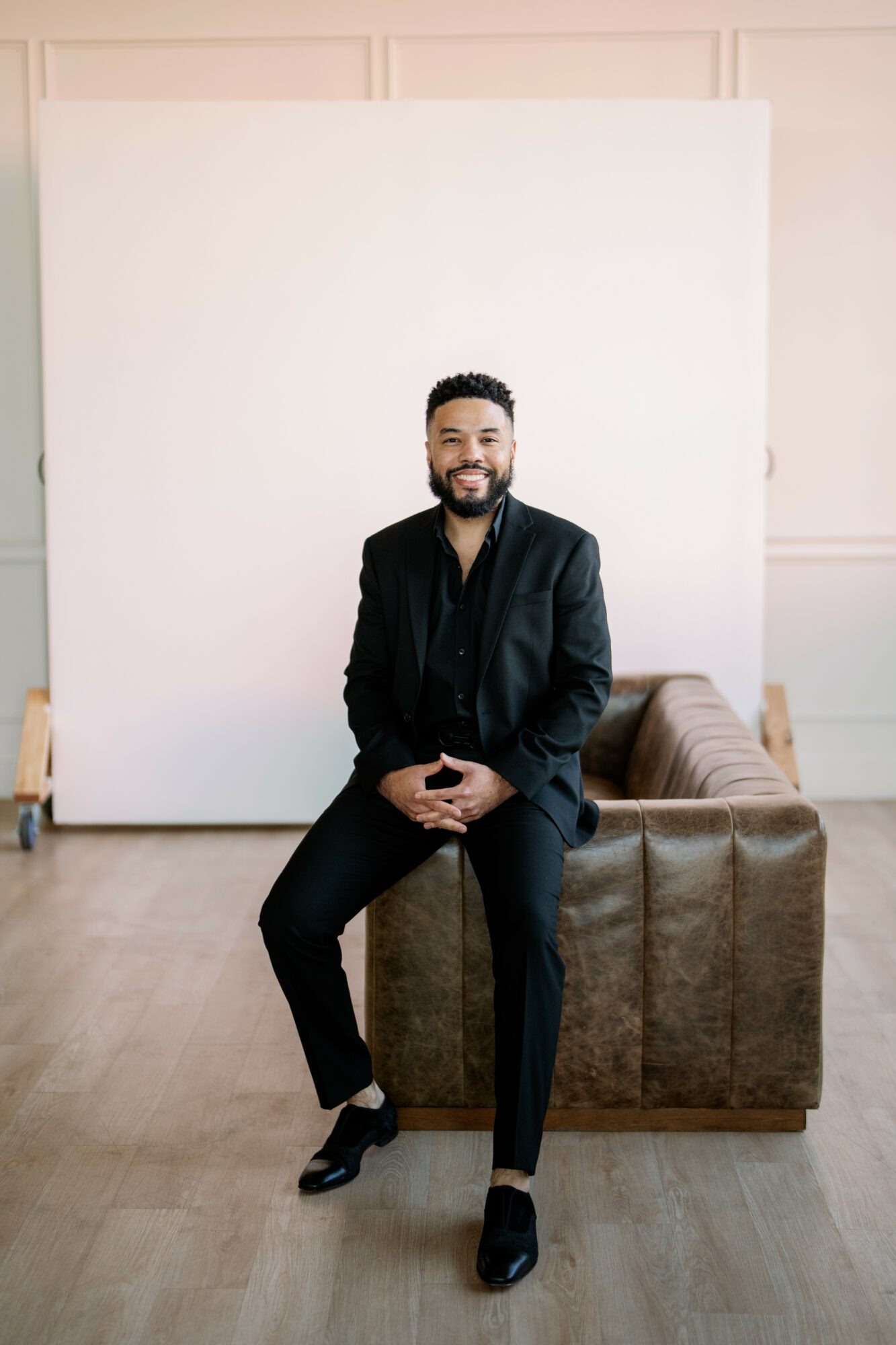Man with a beard in a black suit sitting on a brown leather sofa, smiling, in a studio with a white backdrop.