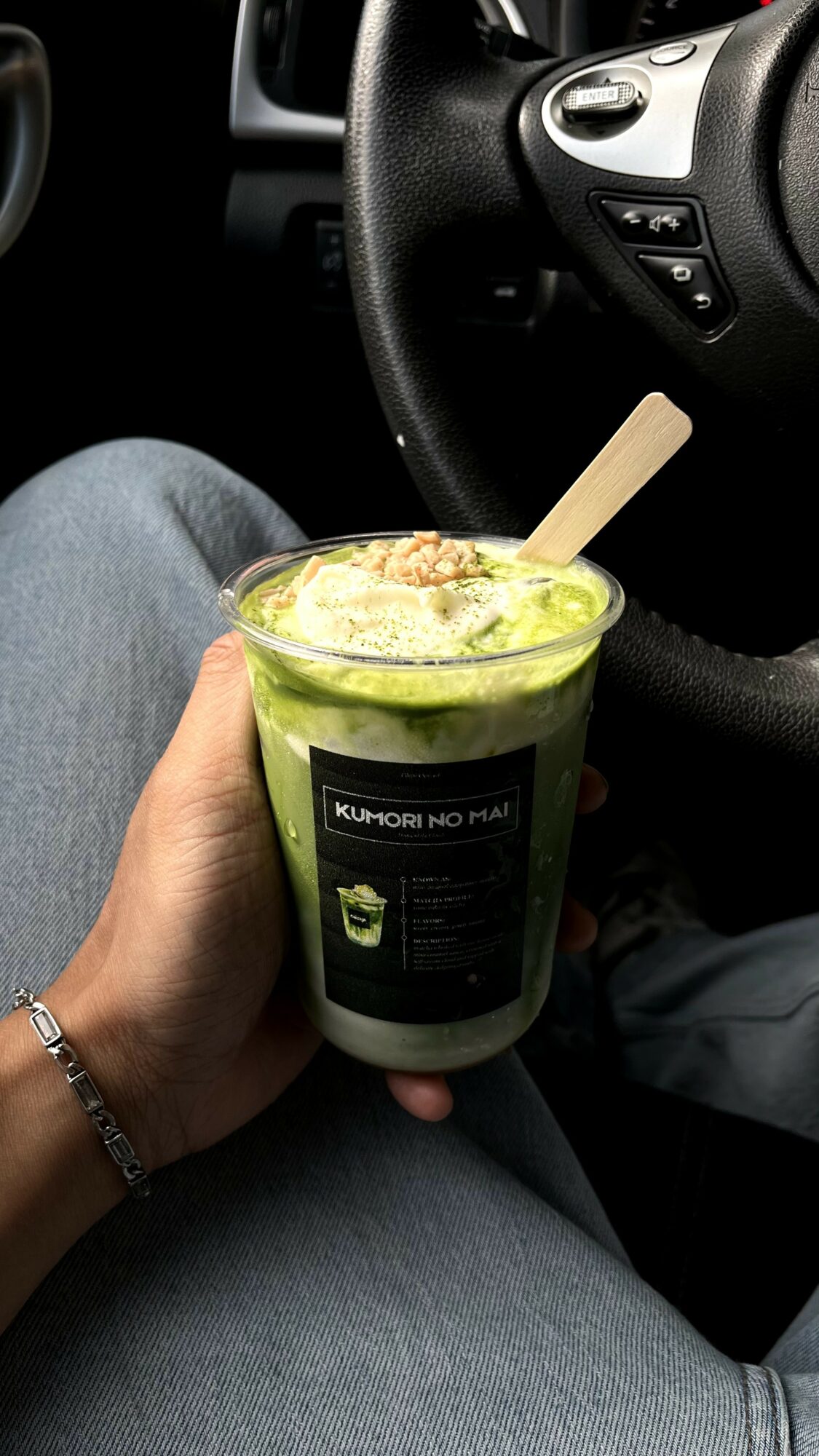Person holding a green smoothie with a wooden spoon inside, sitting in a car with a steering wheel visible.