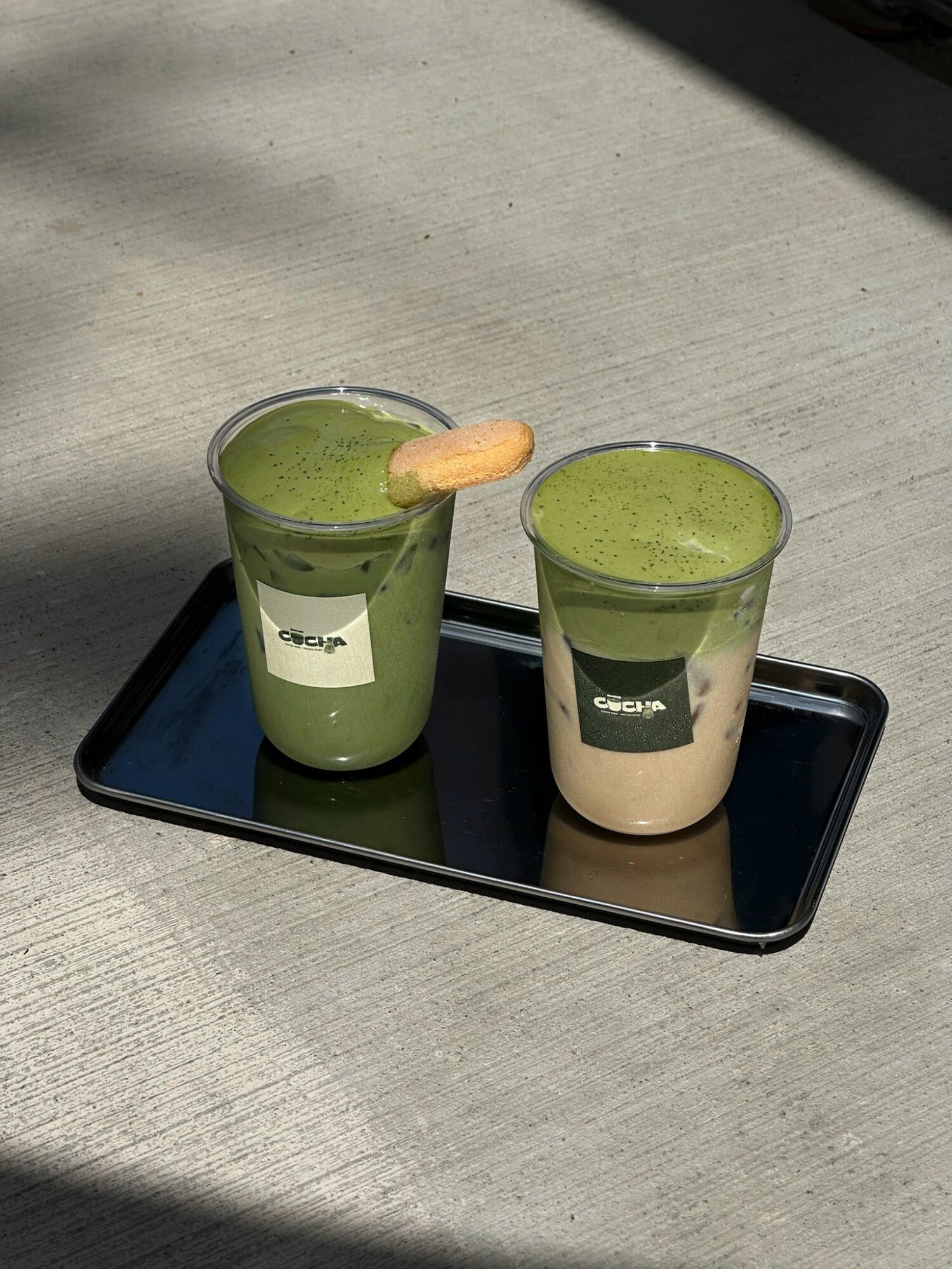 Two glasses of green and beige layered drinks on a black tray, placed on a light gray surface with shadows.