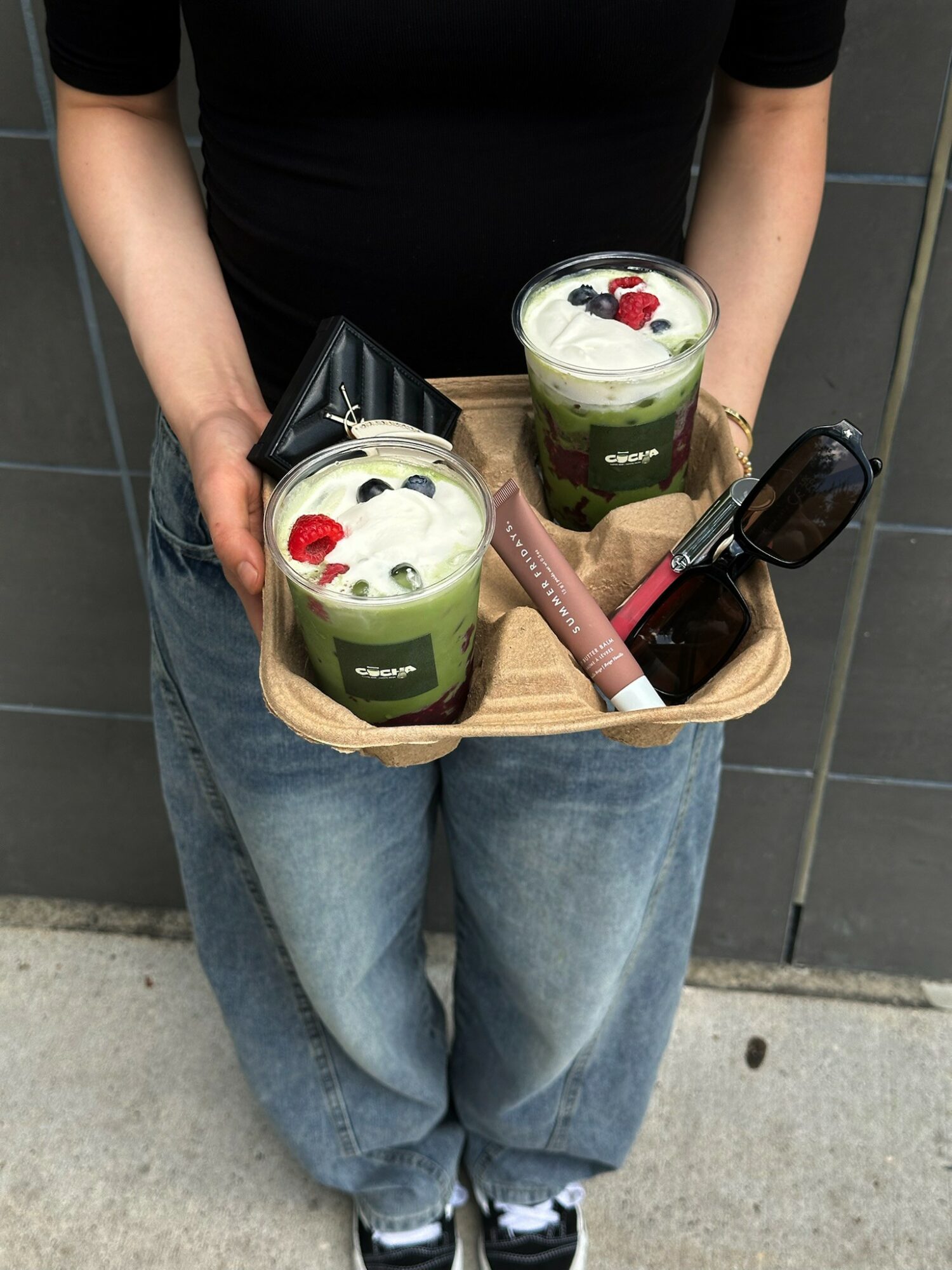 Person holding a tray with two green drinks topped with whipped cream and red berries, a pink item, and a black object.