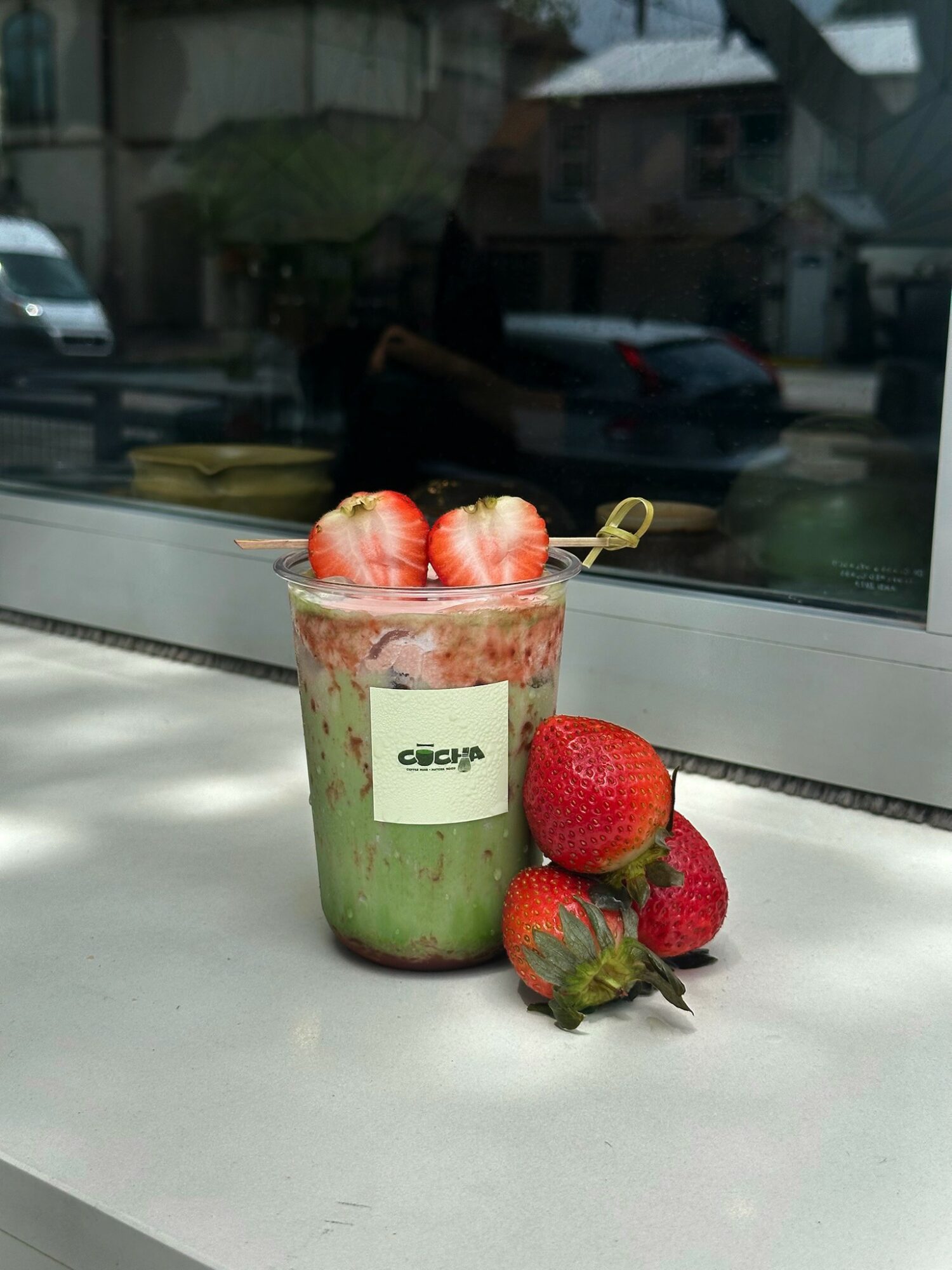 Strawberry and green layered dessert in a glass jar with strawberries beside it on a windowsill.