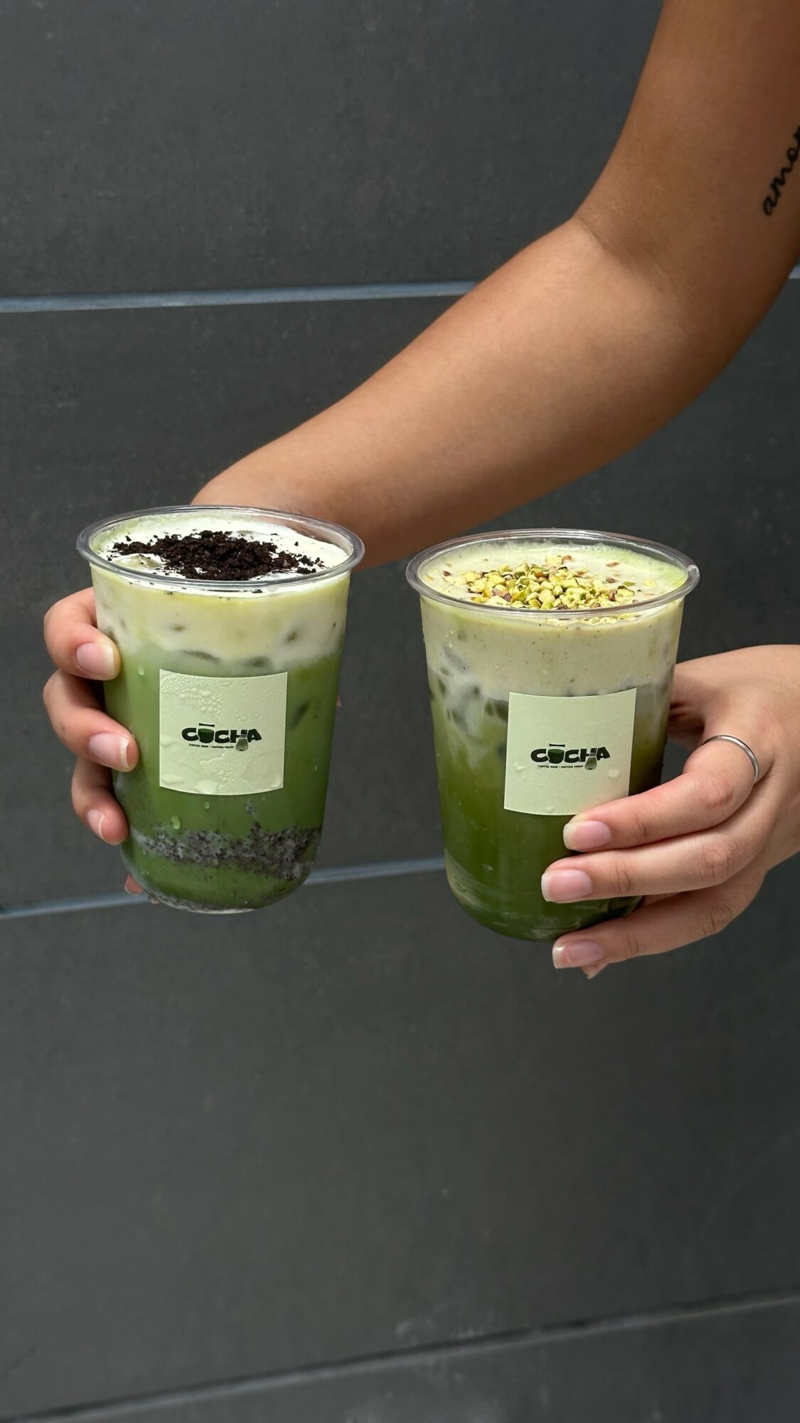 Two hands hold glasses with green layered drinks topped with white foam and sprinkled toppings, against a dark background.