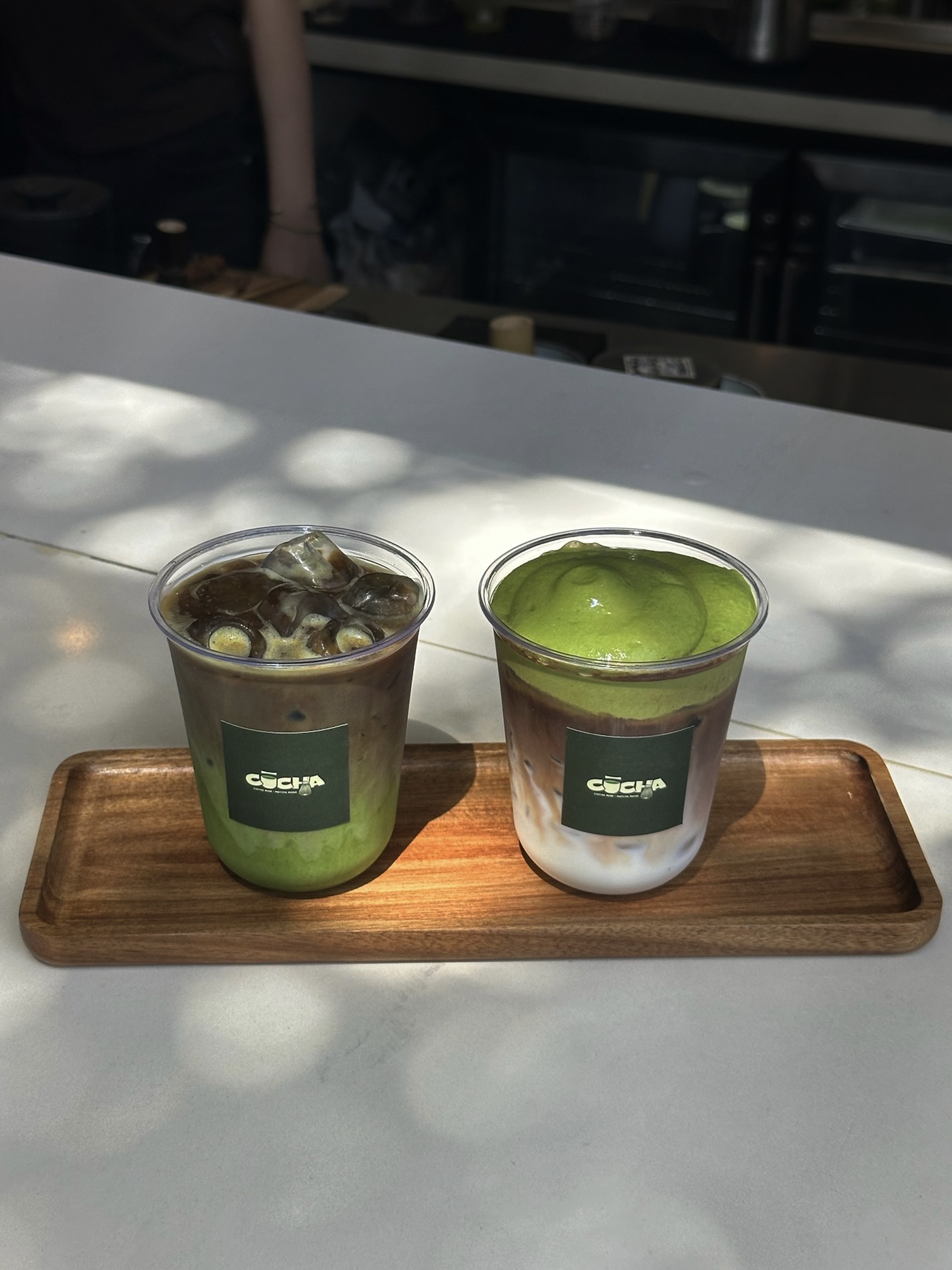 Two cups of green tea on a wooden tray, one with ice and the other with a green fruit, on a white surface.
