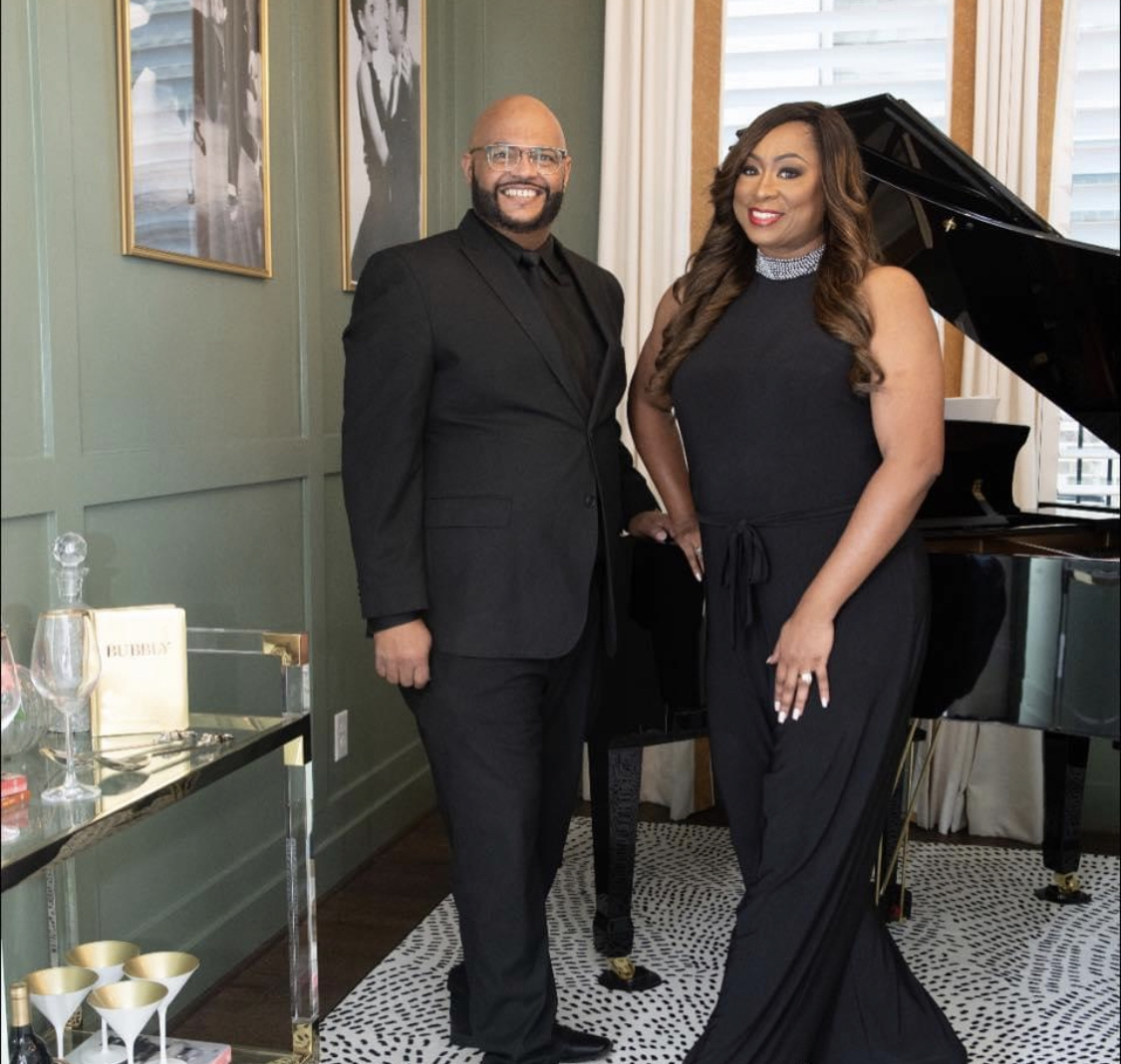 Two people, a man and a woman, stand in a room with a piano, smiling at the camera.