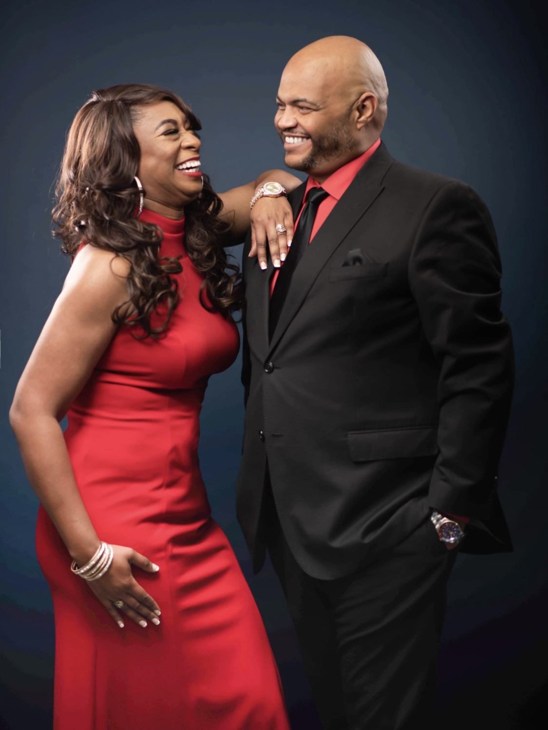 A woman in a red dress and a man in a black suit smile and look at each other, standing close together.