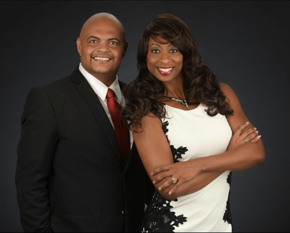 A man and woman in formal attire smiling, standing close together against a dark background.