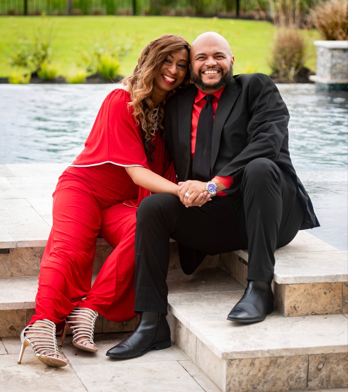 A smiling couple sitting on steps by a pool, woman in red outfit, man in black suit, holding hands, outdoors.