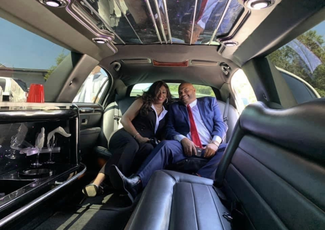 Two people sitting inside a limousine, smiling, with a reflective ceiling and a red siren light visible.