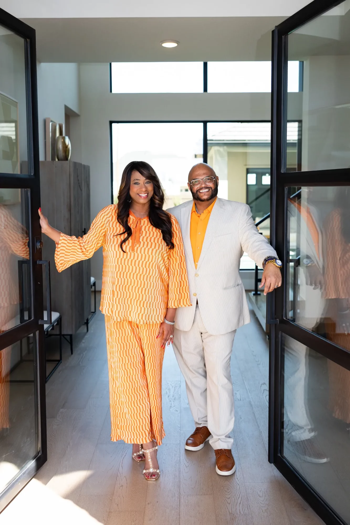 Two people, a woman and a man, stand in a modern indoor space with large windows, smiling and posing together.