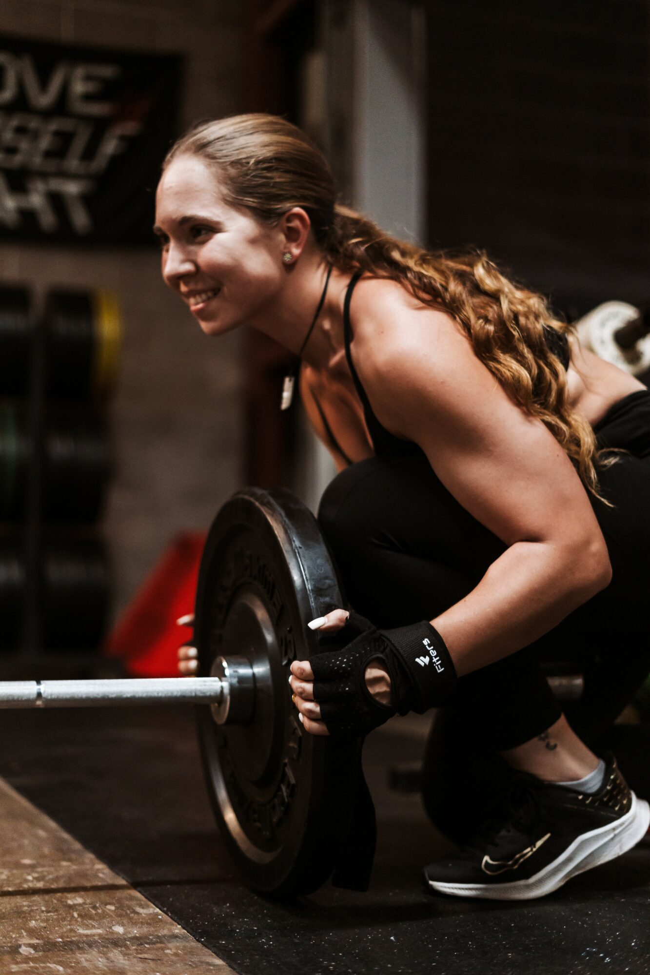 Woman lifting a barbell in a gym, smiling, with long hair tied back, wearing workout clothes and gloves.