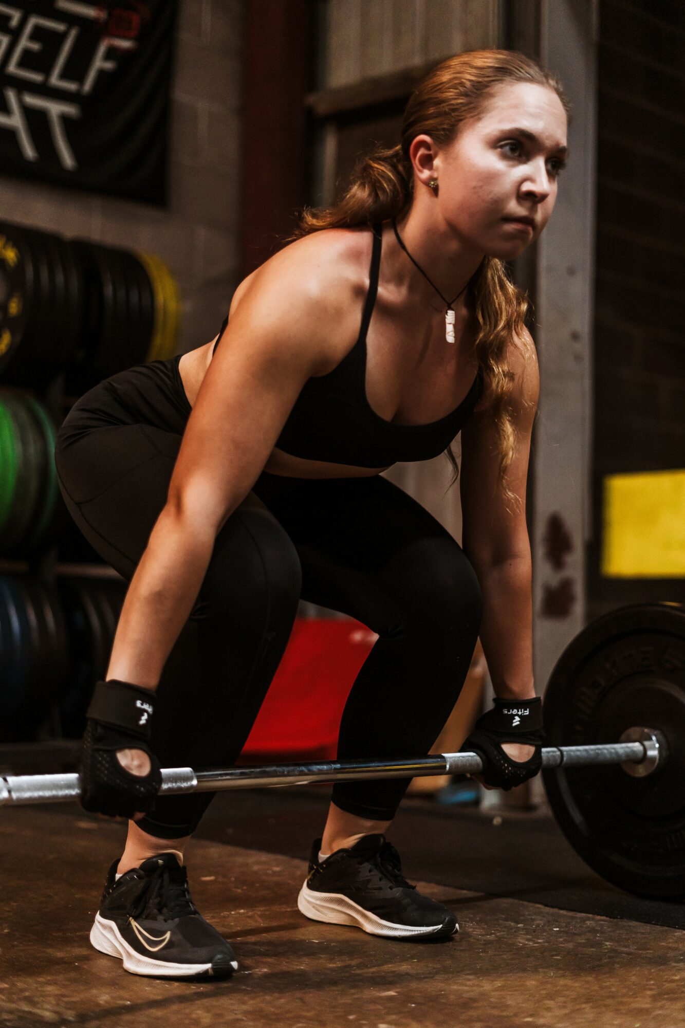 Woman with red hair in workout clothes lifting a barbell in a gym, focused expression, dark background.