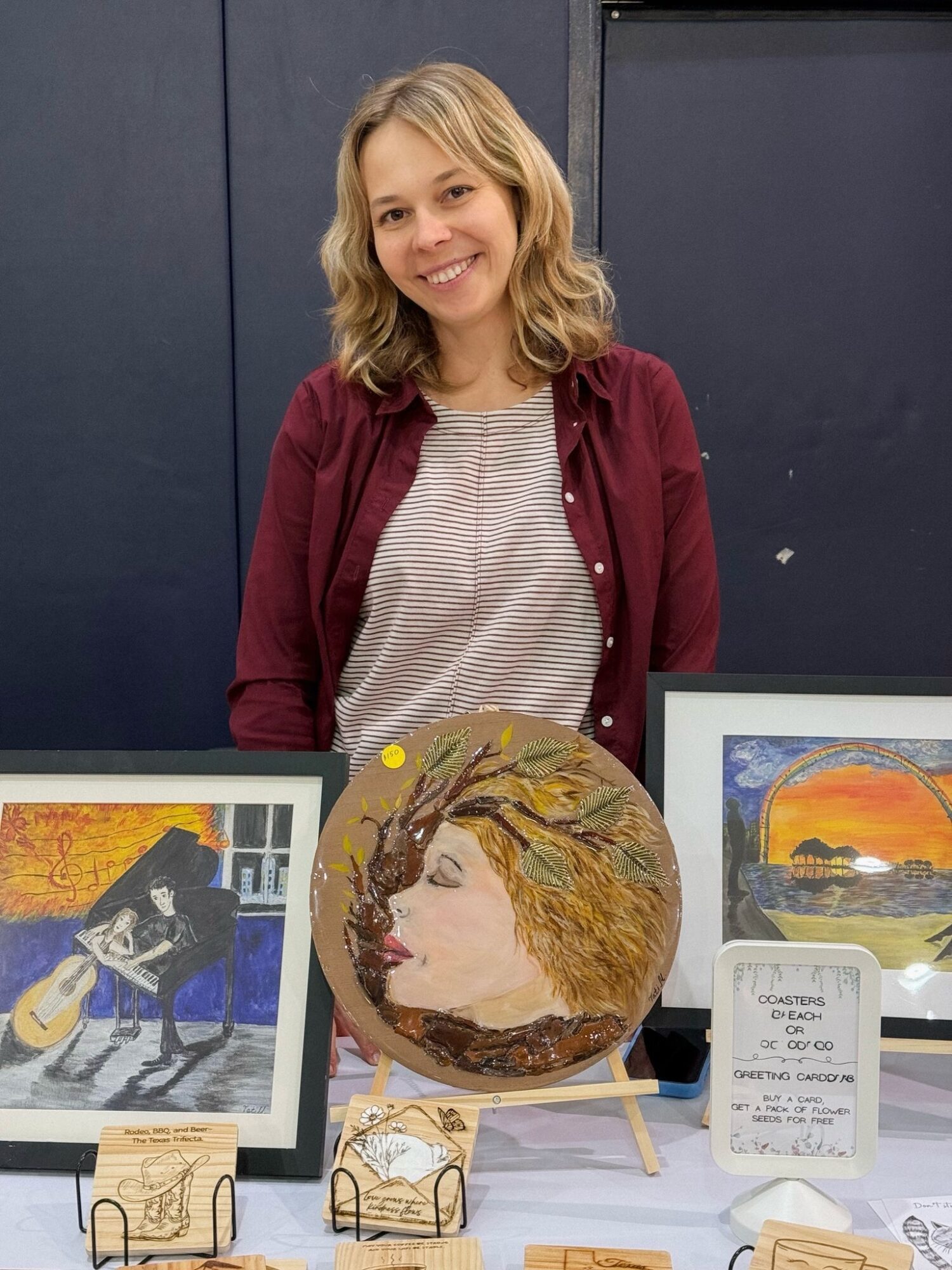 Young woman with blonde hair smiling, standing behind artwork including a painted face on a round object, in an indoor setting.