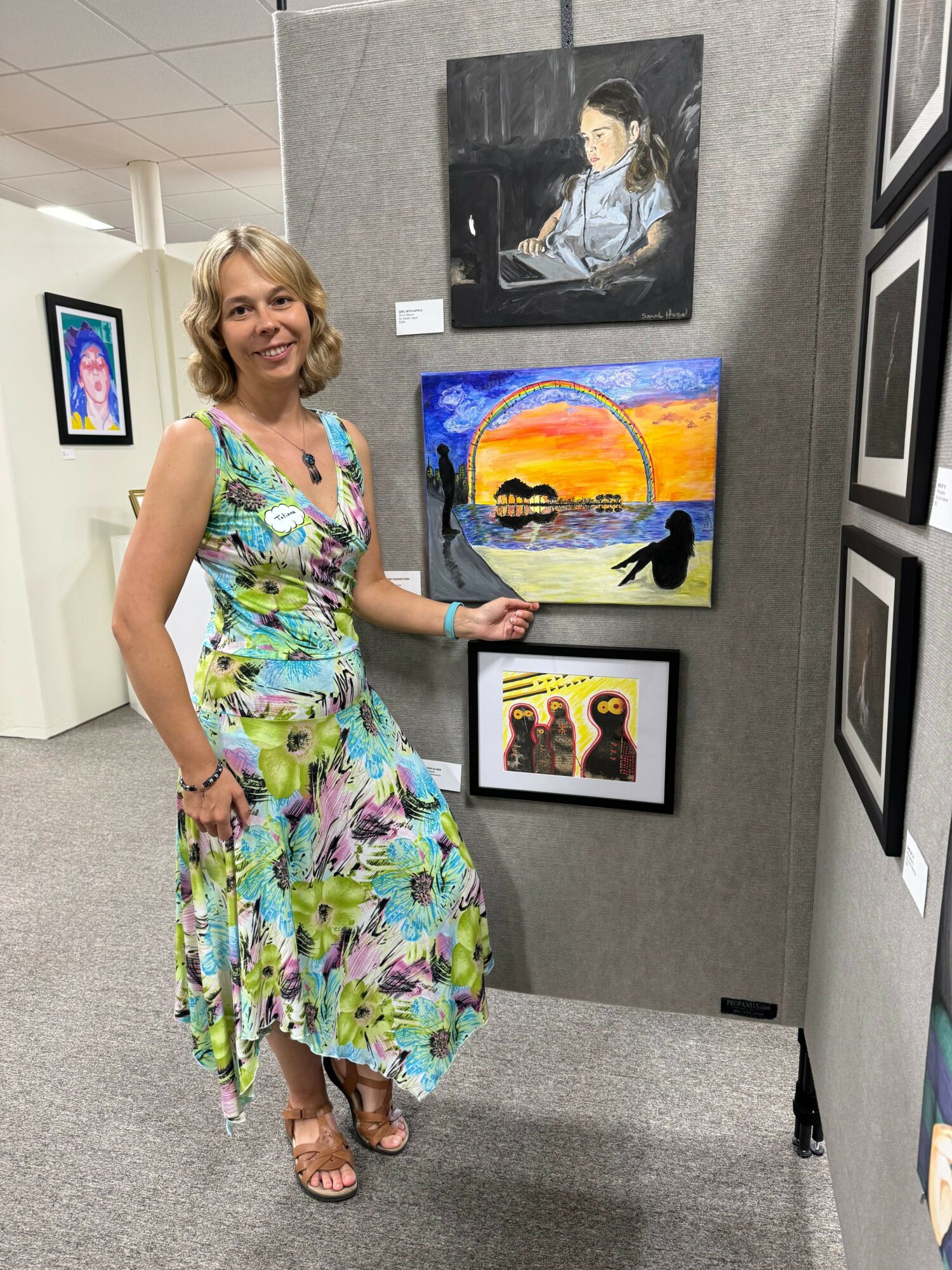 Woman in a colorful floral dress stands in an art gallery, pointing at paintings on the wall.