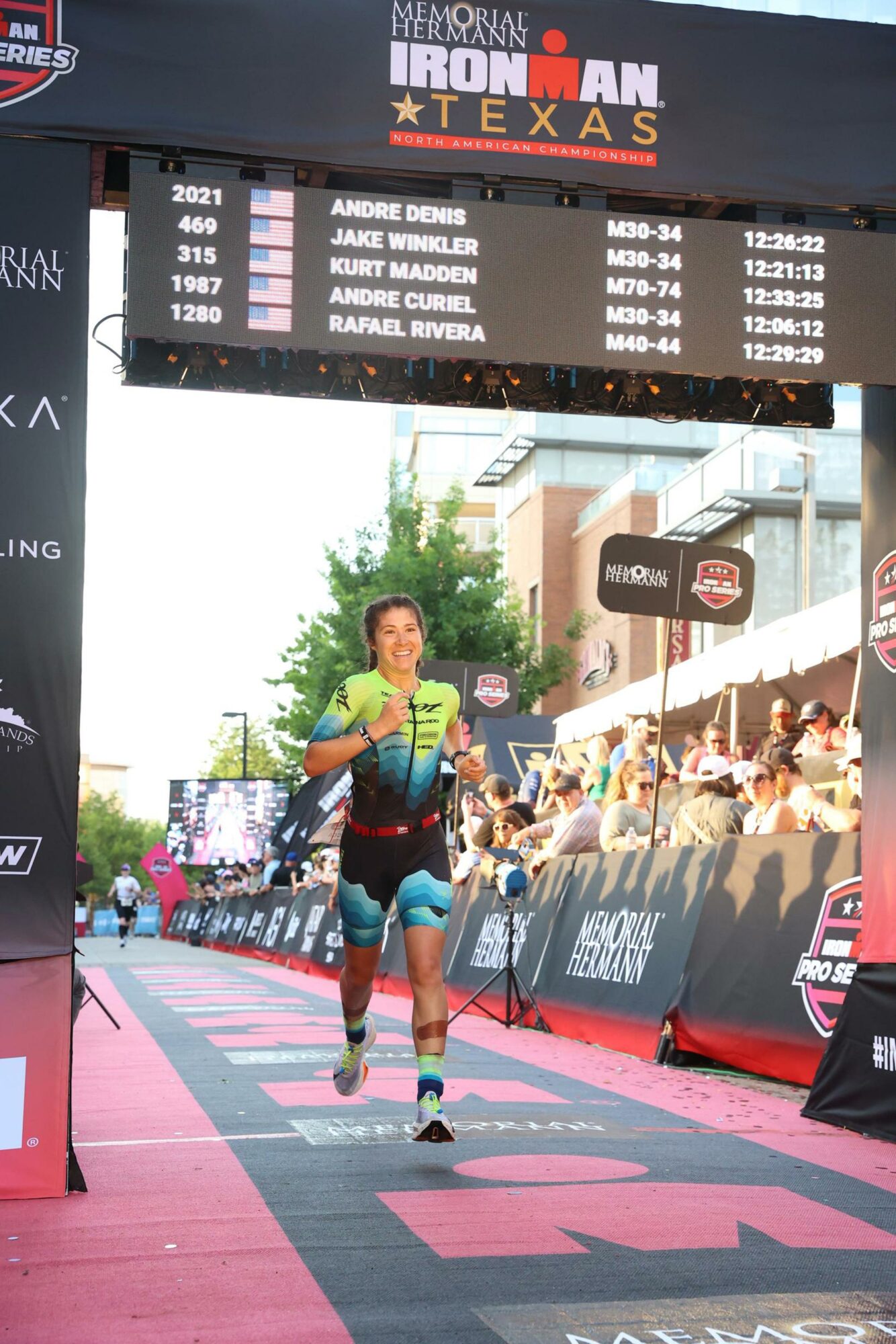 Female athlete crossing finish line at Ironman Texas race, with spectators and event banners in background.