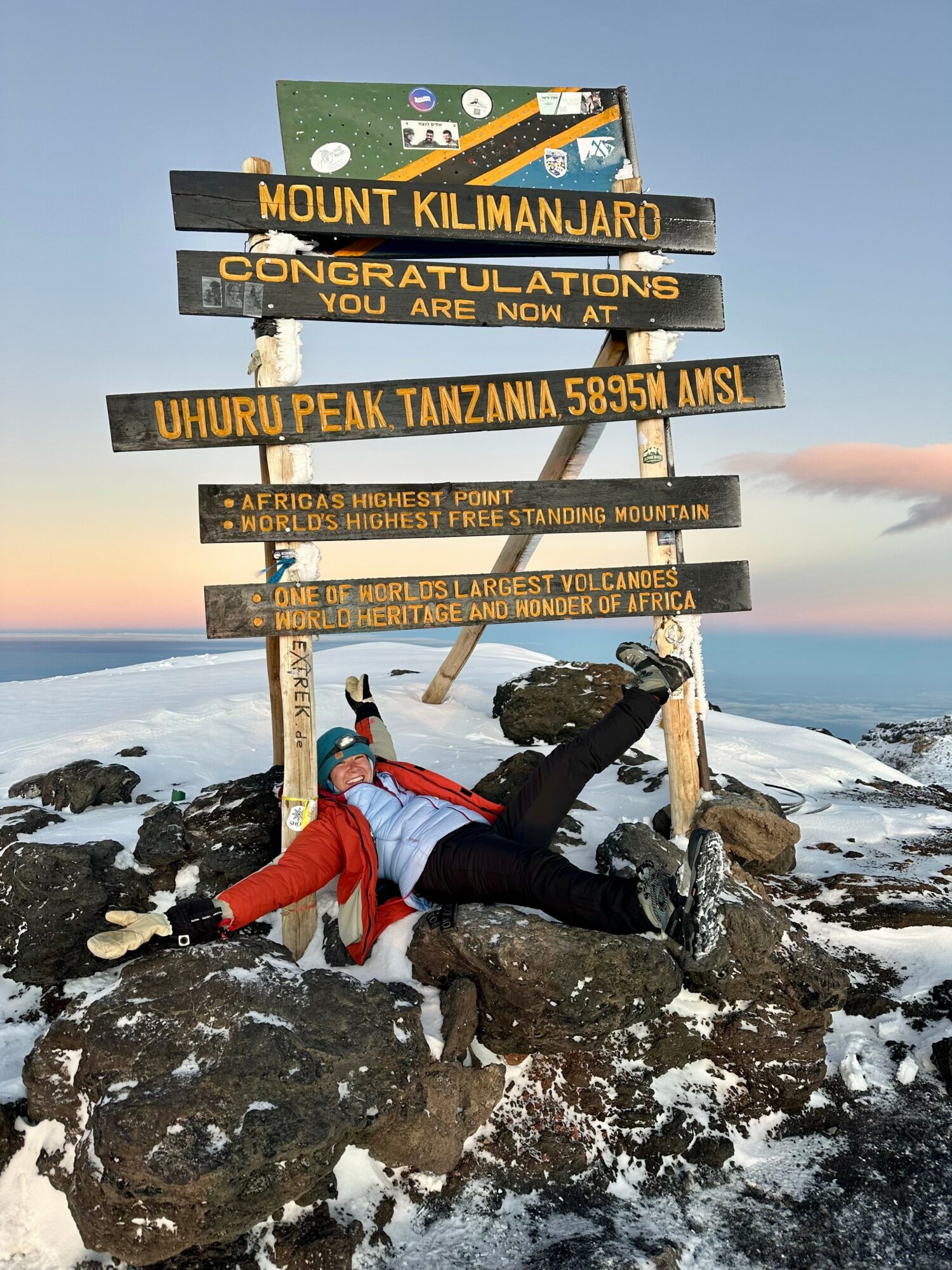 Person lying on rocks with snow, wearing red and black, next to a signpost at Mount Kilimanjaro summit, with a mountain and sky background.