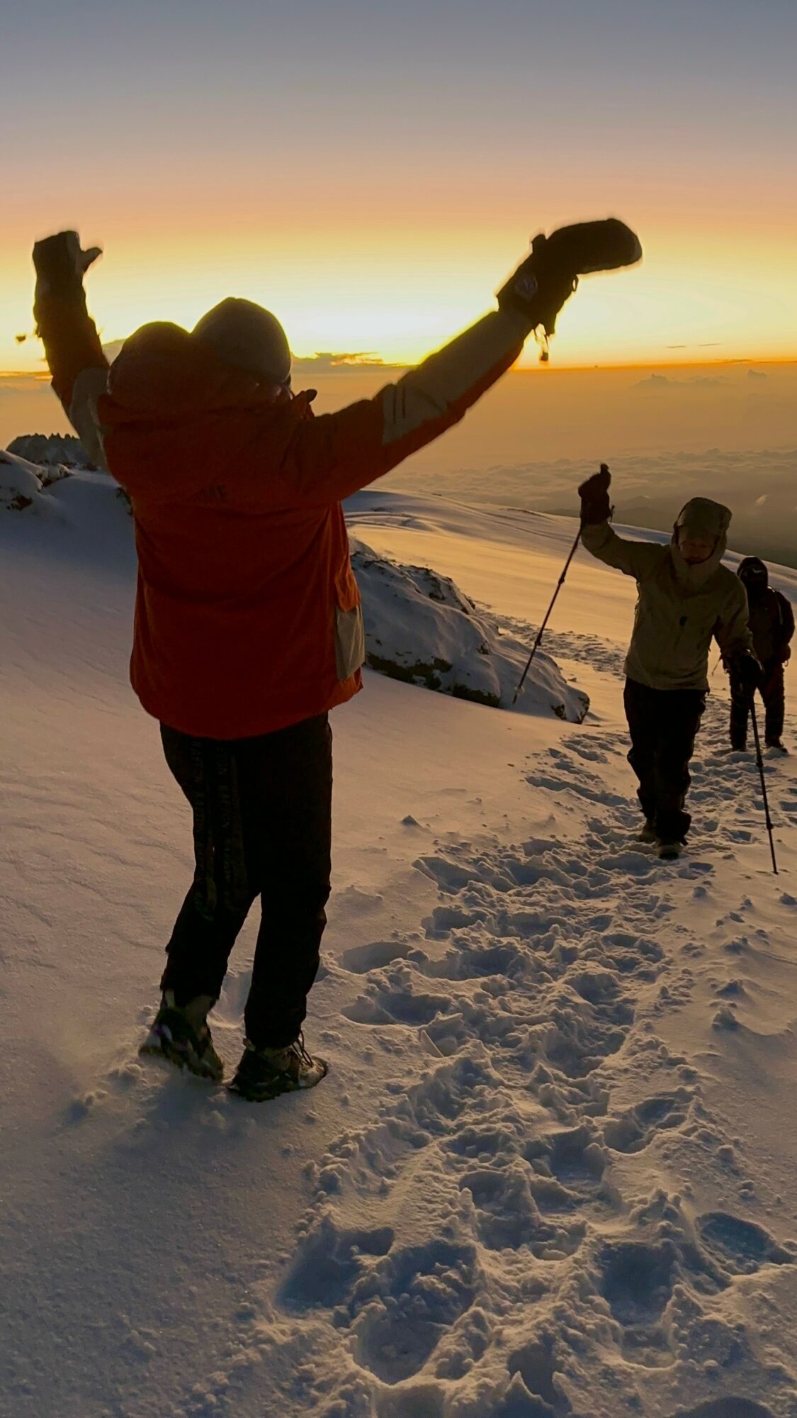 Group of people hiking on snow-covered terrain during sunset, some raising arms, with a mountain landscape in the background.