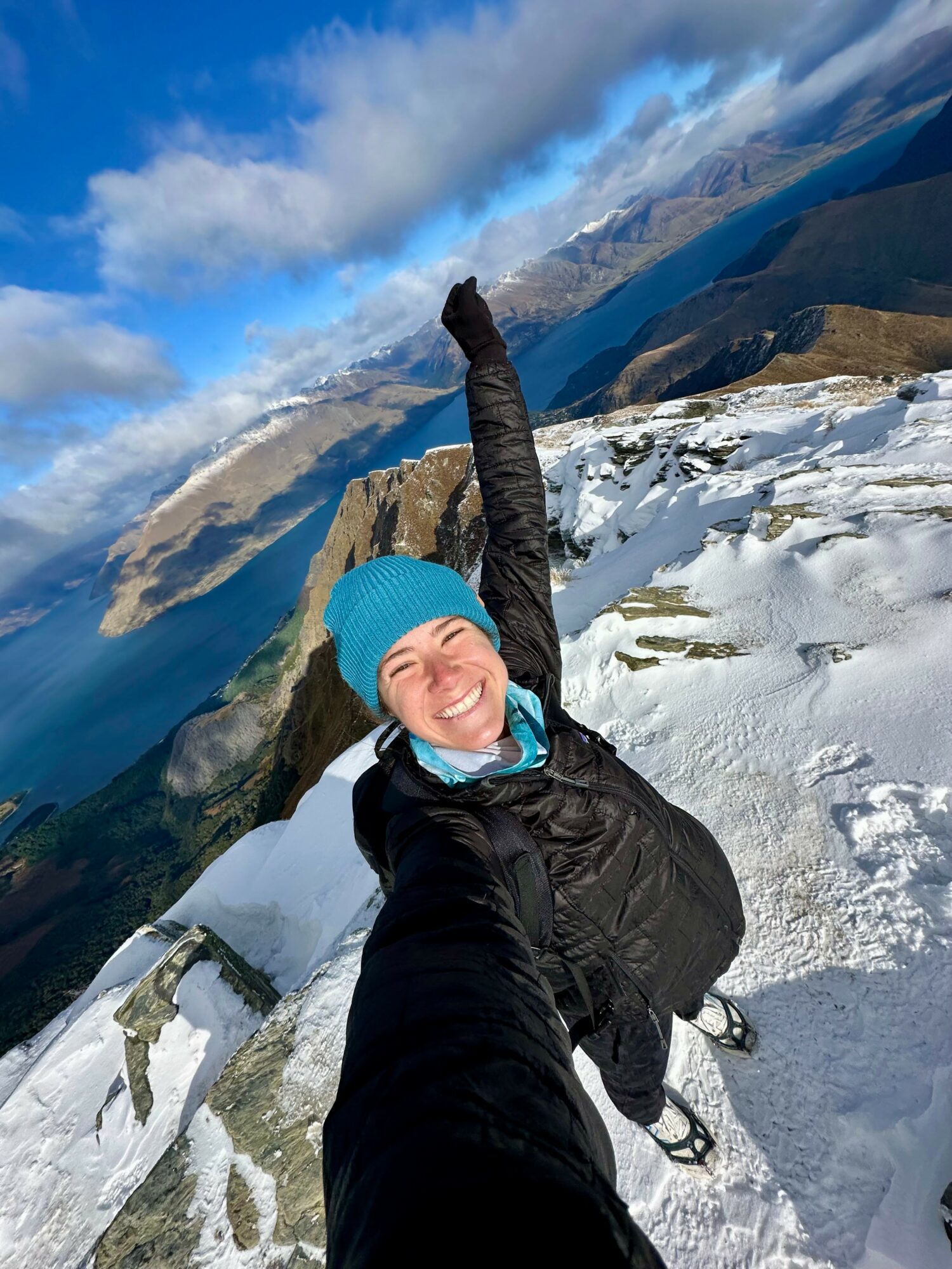 Person smiling, wearing a blue beanie and black jacket, taking a selfie on snowy mountain slope with clouds and mountains in background.