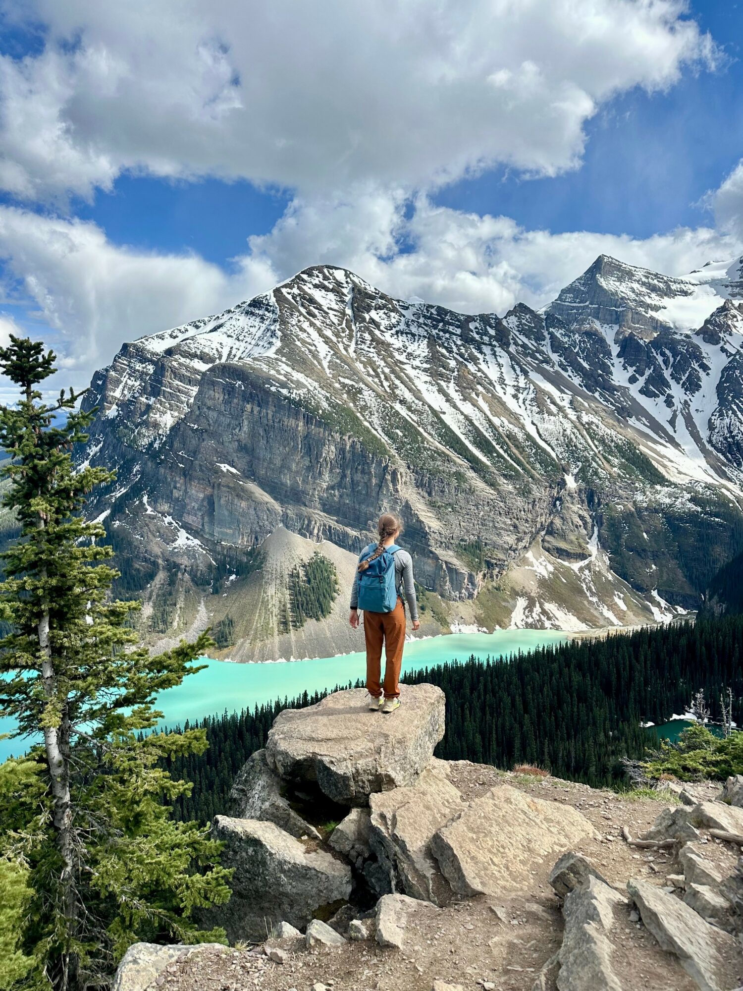 Person standing on rocks overlooking a turquoise lake with snow-capped mountains and a partly cloudy sky.