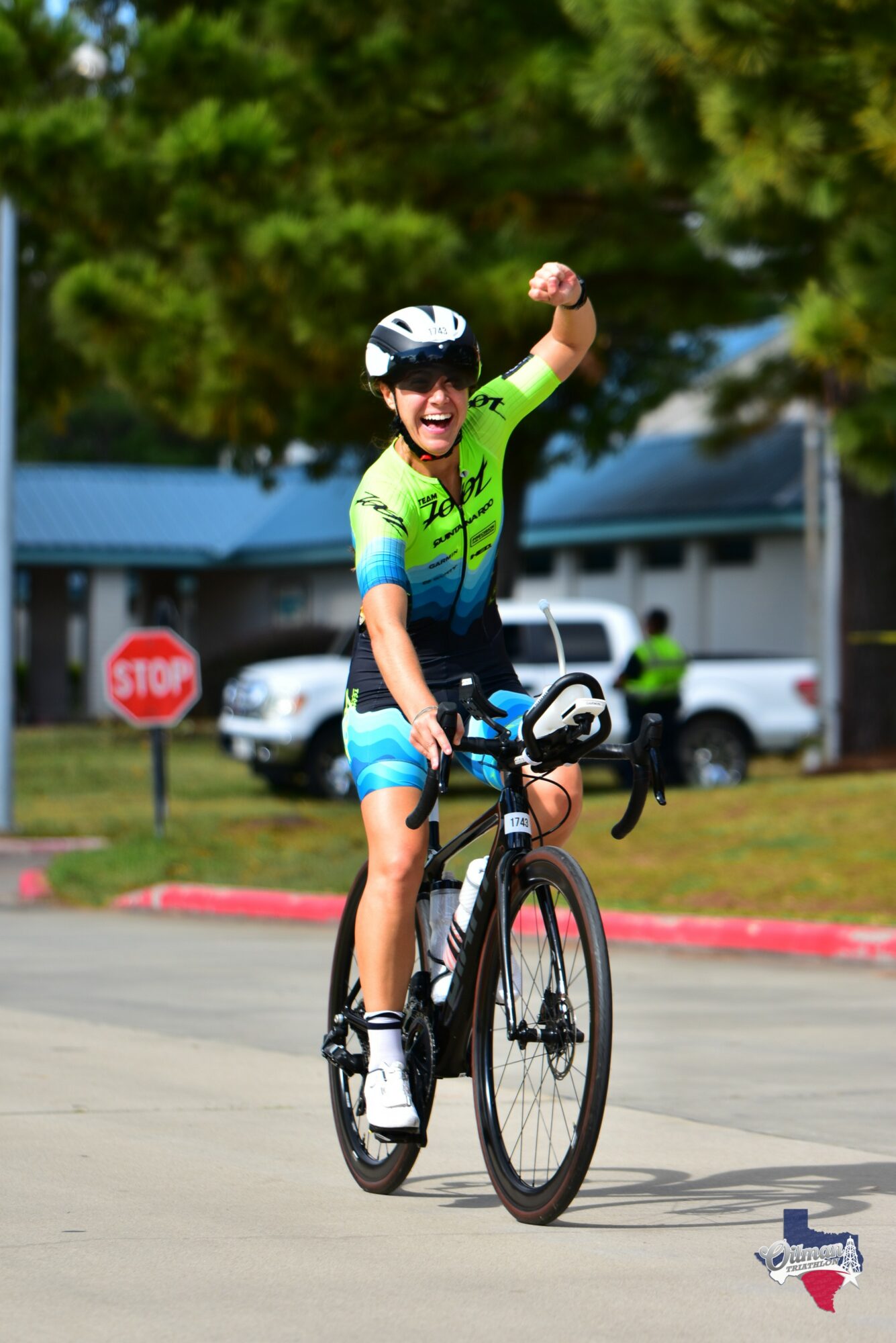 Child riding a bicycle with one arm raised, wearing a helmet and sunglasses, in a park-like setting.