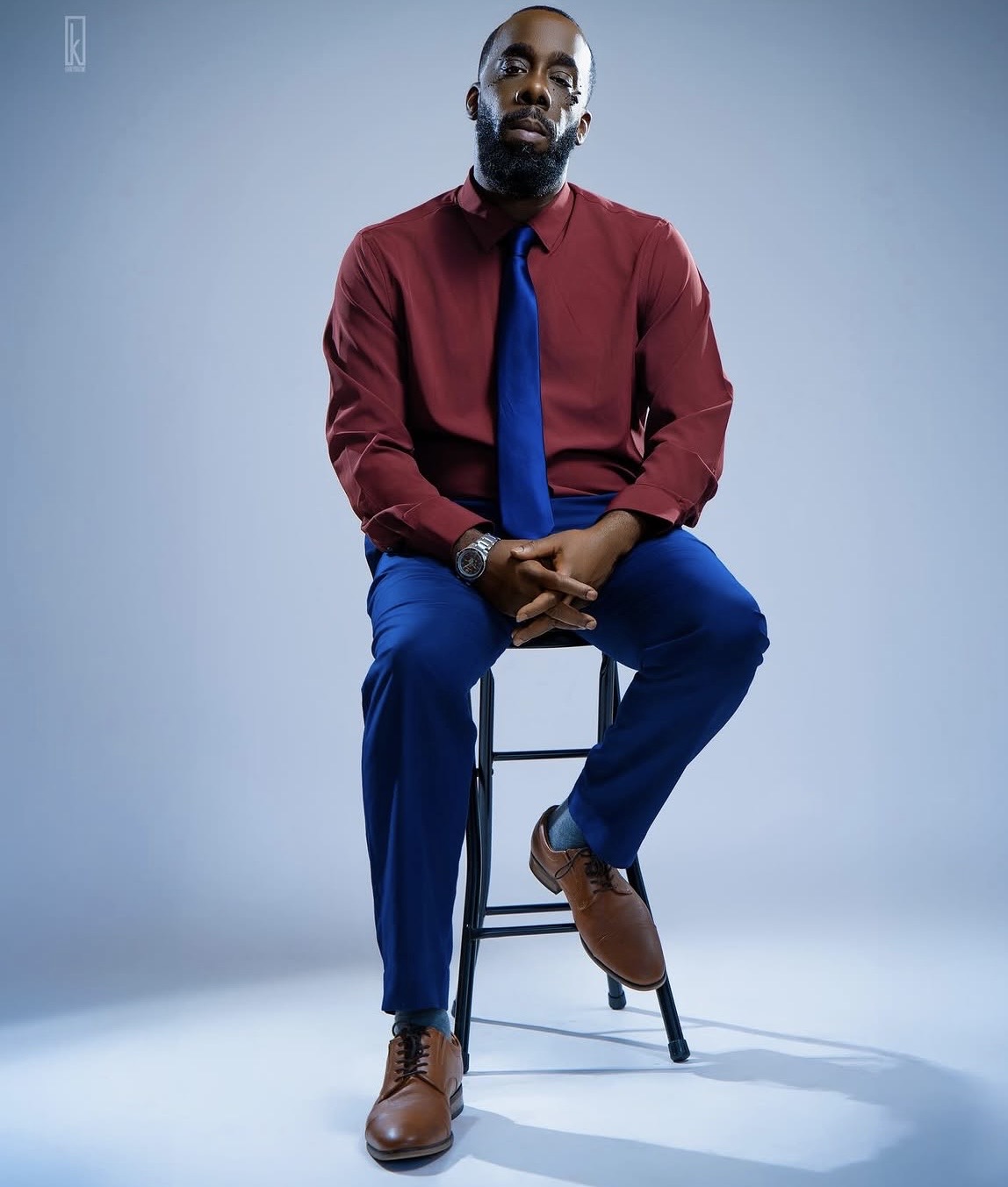 Man sitting on a stool, wearing a red shirt, blue pants, and brown shoes, with a blue tie, against a plain background.