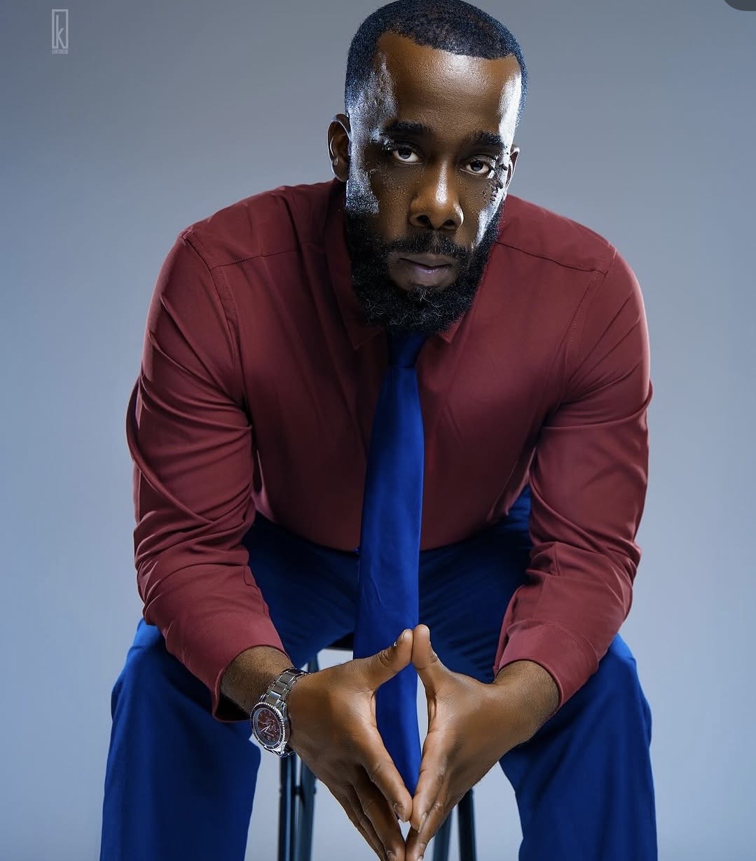 Man in red shirt and blue tie leaning forward with hands clasped, looking at camera, gray background.