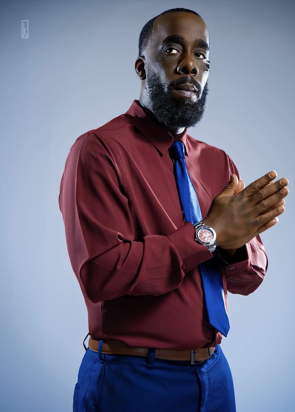 Man with a beard wearing a red shirt, blue tie, and blue pants, holding his hands together in front of him.