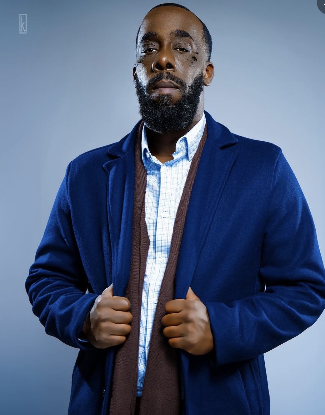 Man with beard in blue jacket and white shirt, holding lapels, against a plain background.