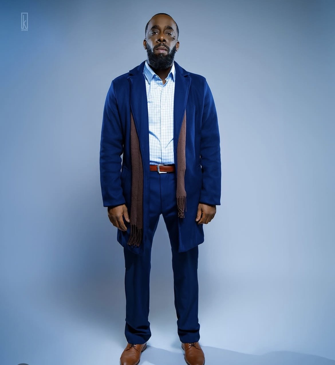 Man in a blue suit and coat standing against a plain background, looking at the camera.