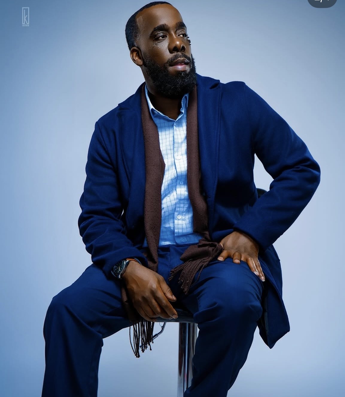 Man with beard sitting on a stool, wearing a blue jacket and shirt, against a light blue background.