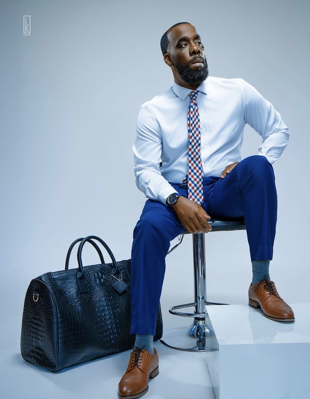 Man sitting on a stool with a large black bag beside him, wearing a light blue shirt, patterned tie, blue pants, and brown shoes.