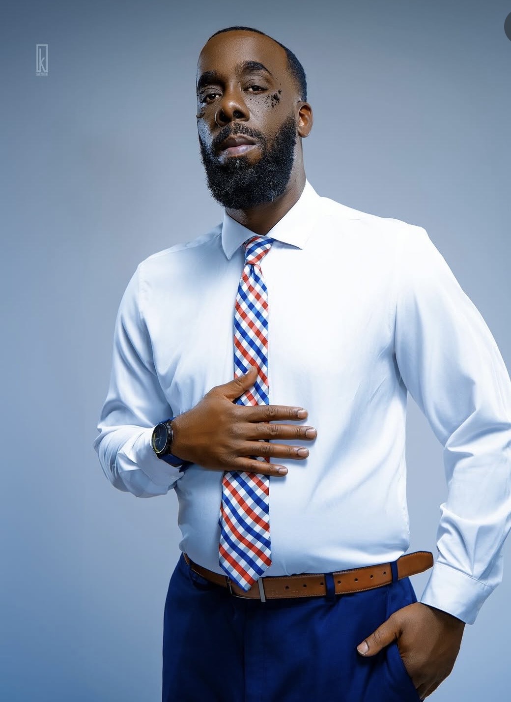 Man in white shirt and colorful tie, standing with hand on chest, against plain background.