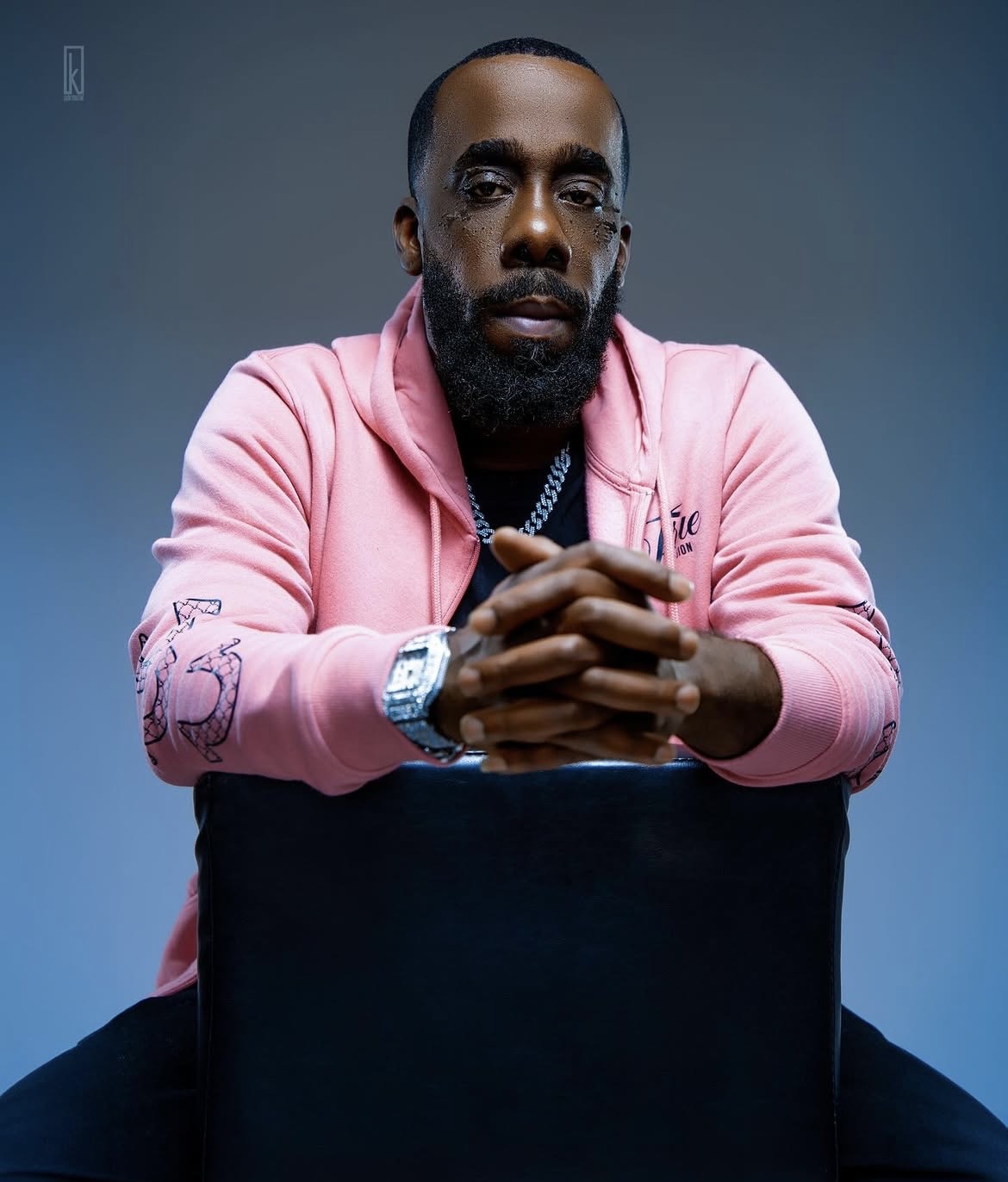 Man with a beard and glasses wearing a pink hoodie, sitting with hands clasped, against a plain background.