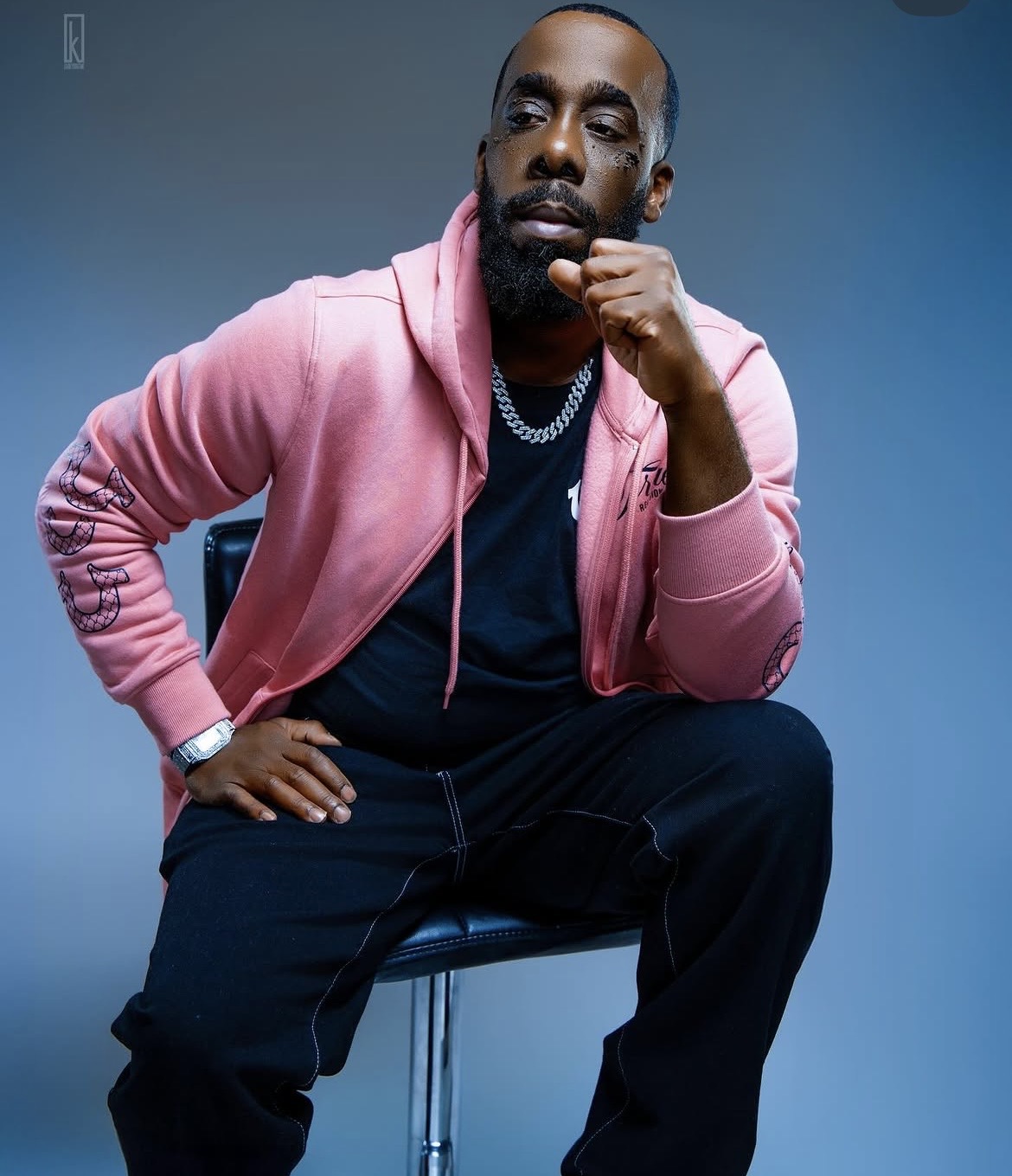 Man sitting with hand on chin, wearing pink hoodie, black pants, and jewelry, against a blue background.