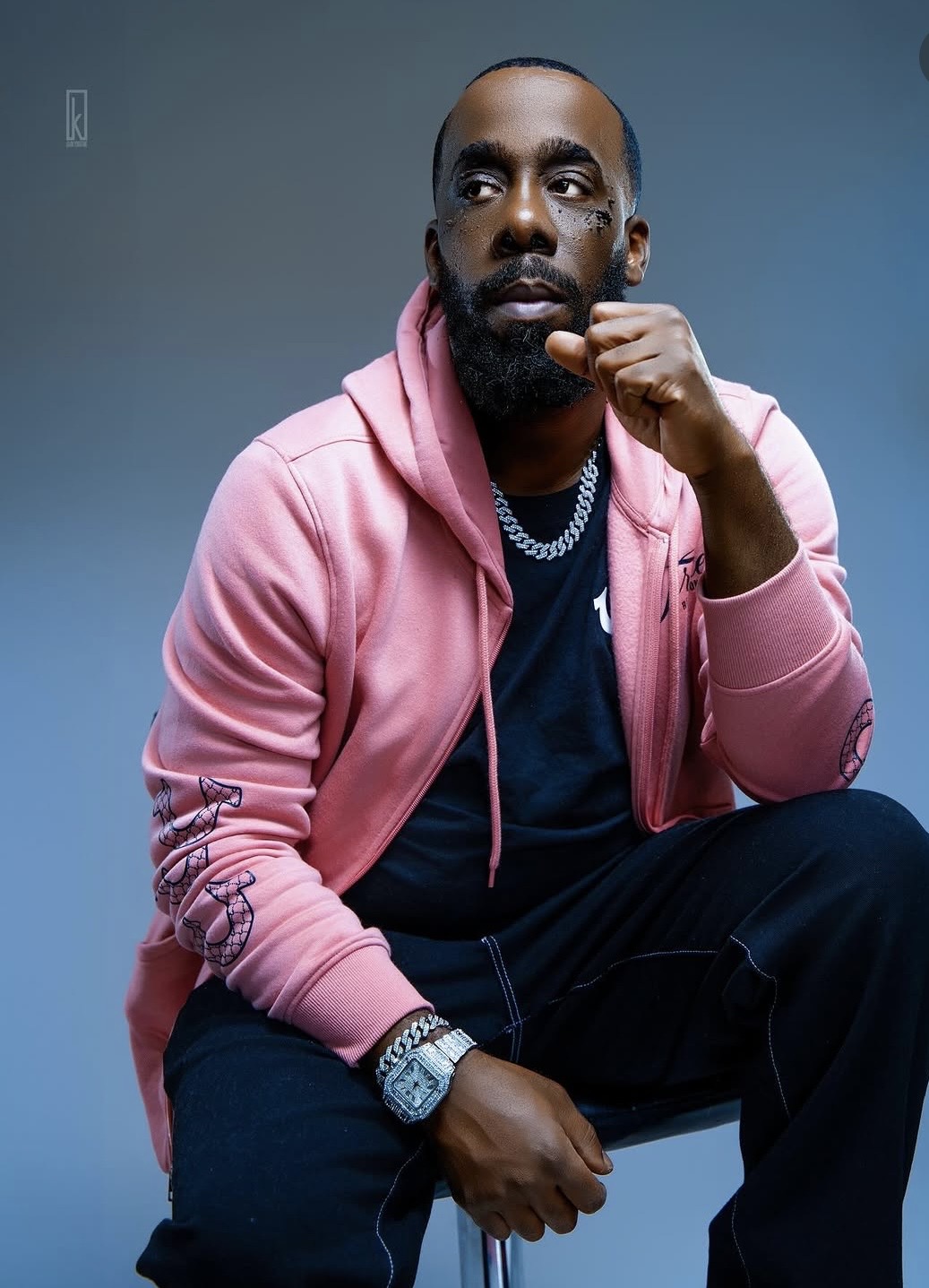 Man with a beard wearing a pink hoodie, sitting against a blue background, looking to the side, with a watch and chain.