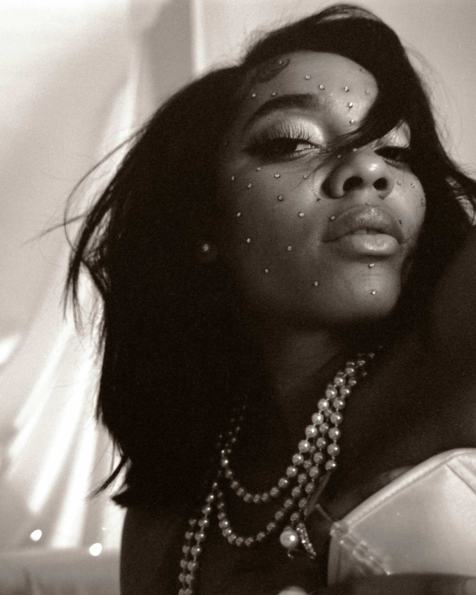 Woman with dark hair, adorned with pearls and rhinestones on face, looking at camera with closed eyes, in black and white.
