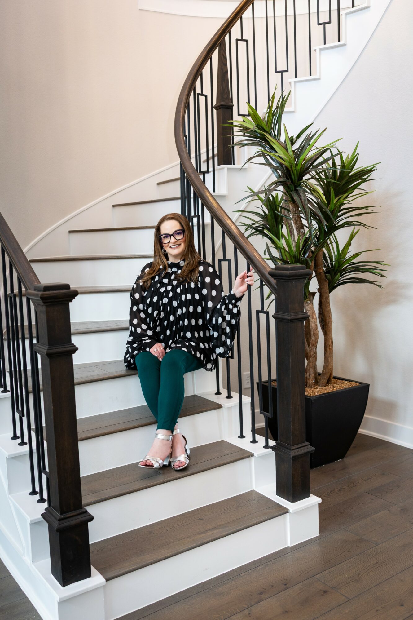 Woman sitting on staircase with black and white polka dot top, teal pants, and pink shoes, next to potted plant.