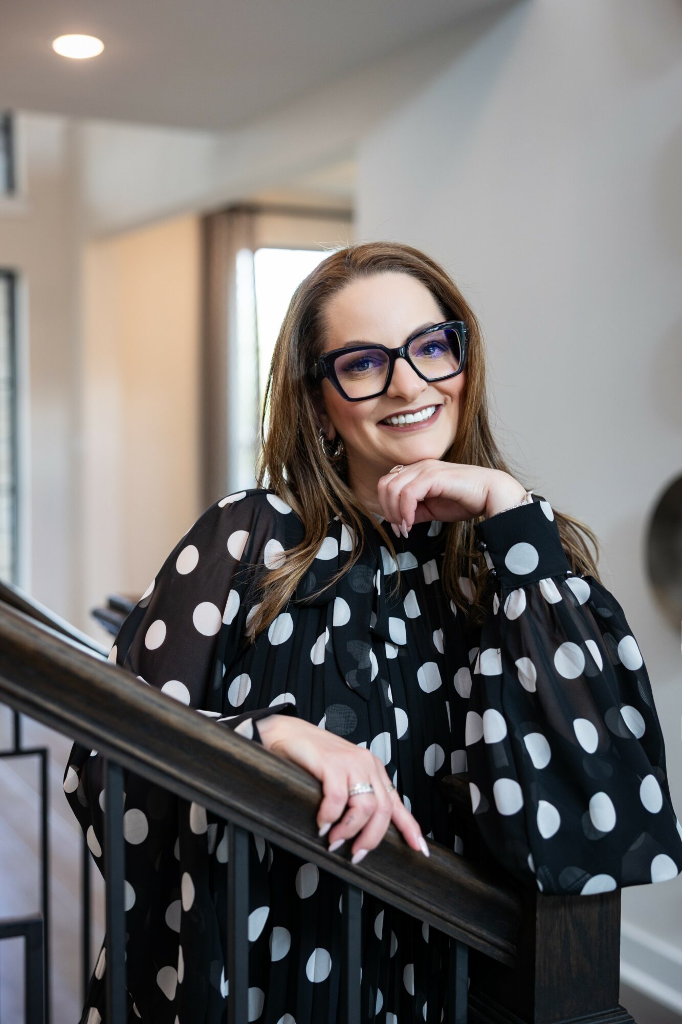 Woman with glasses smiling, wearing a black and white polka dot blouse, leaning on a staircase railing indoors.