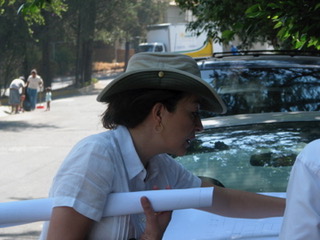 Woman wearing a hat and white shirt leaning on a white railing outdoors, with trees and vehicles in background.