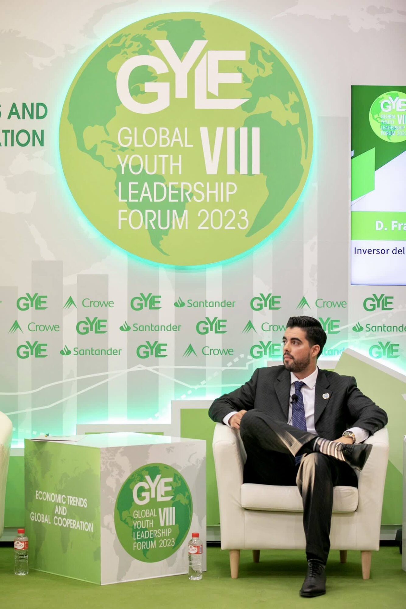 Man in suit sitting on a white chair on stage with green and white backdrop and GYE logo, at youth leadership forum.