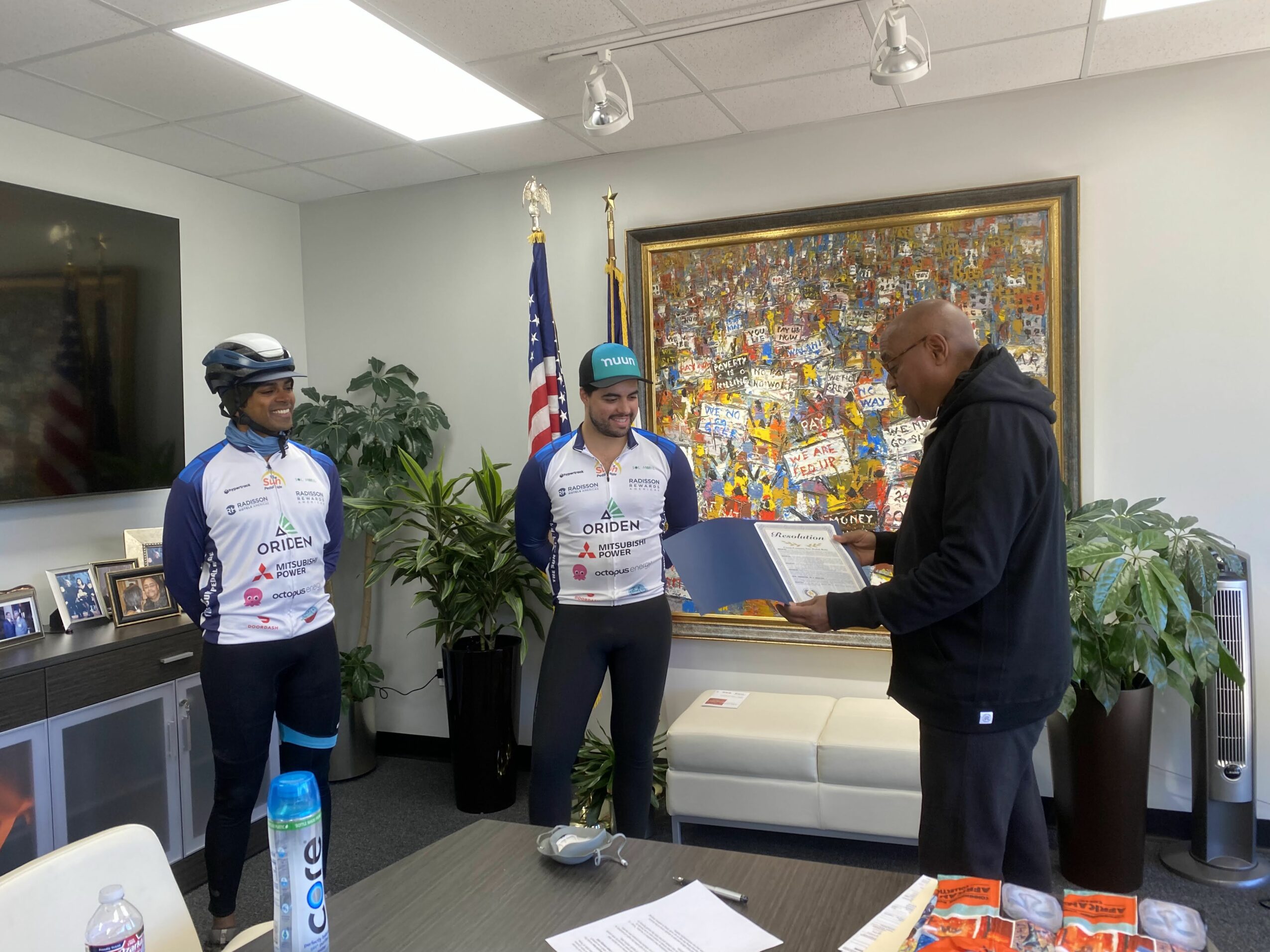 Three people in cycling jerseys and a man in a black hoodie in an office with plants, a painting, and American flags.