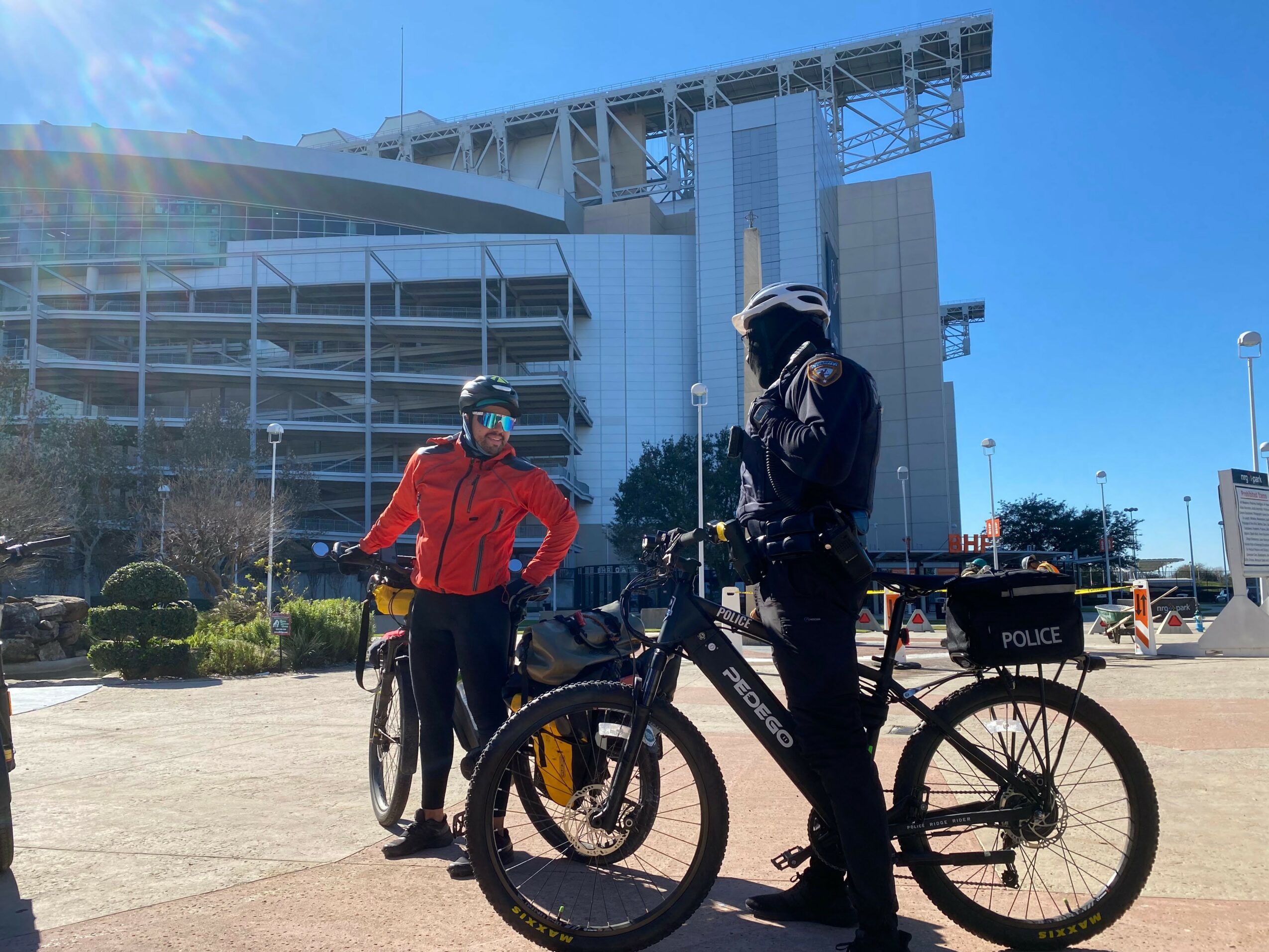 Two people with bicycles stand outdoors near a modern building, one in a red jacket and the other in black, both wearing helmets.