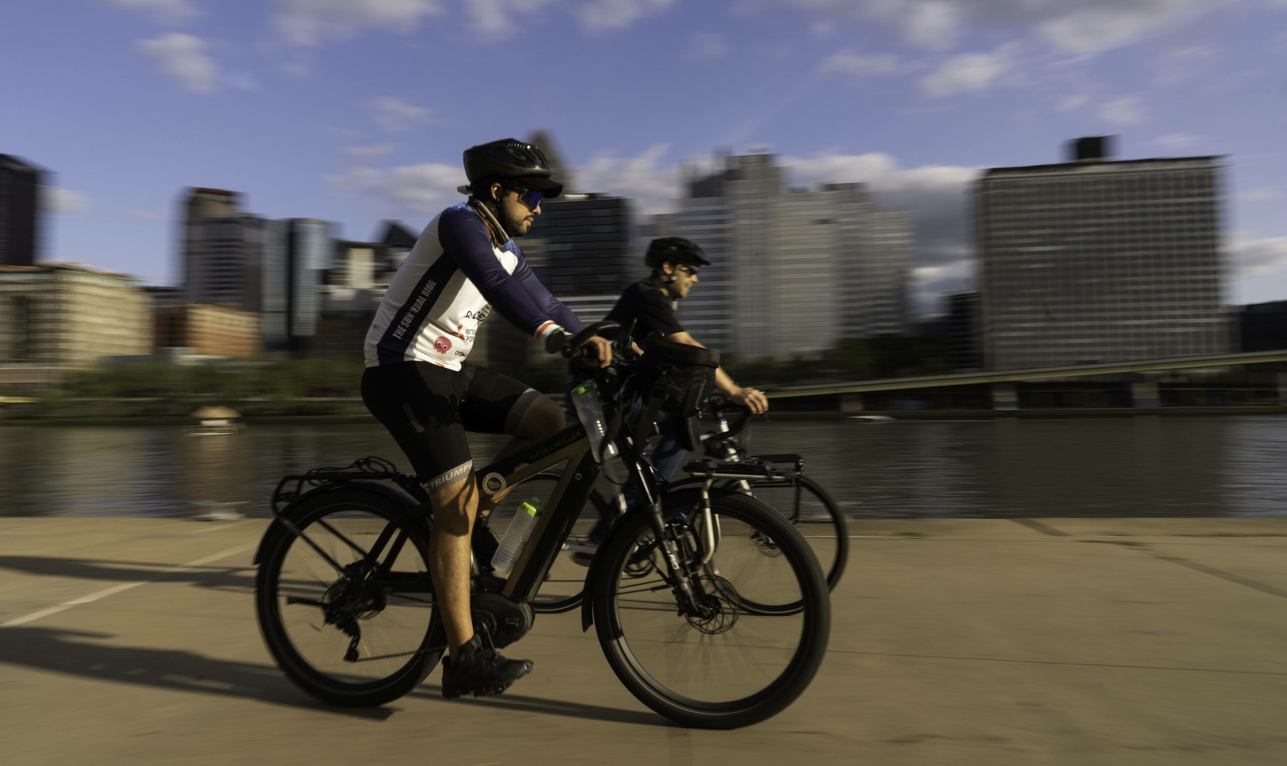 Two people riding bicycles along a city waterfront with tall buildings in the background, under a partly cloudy sky.