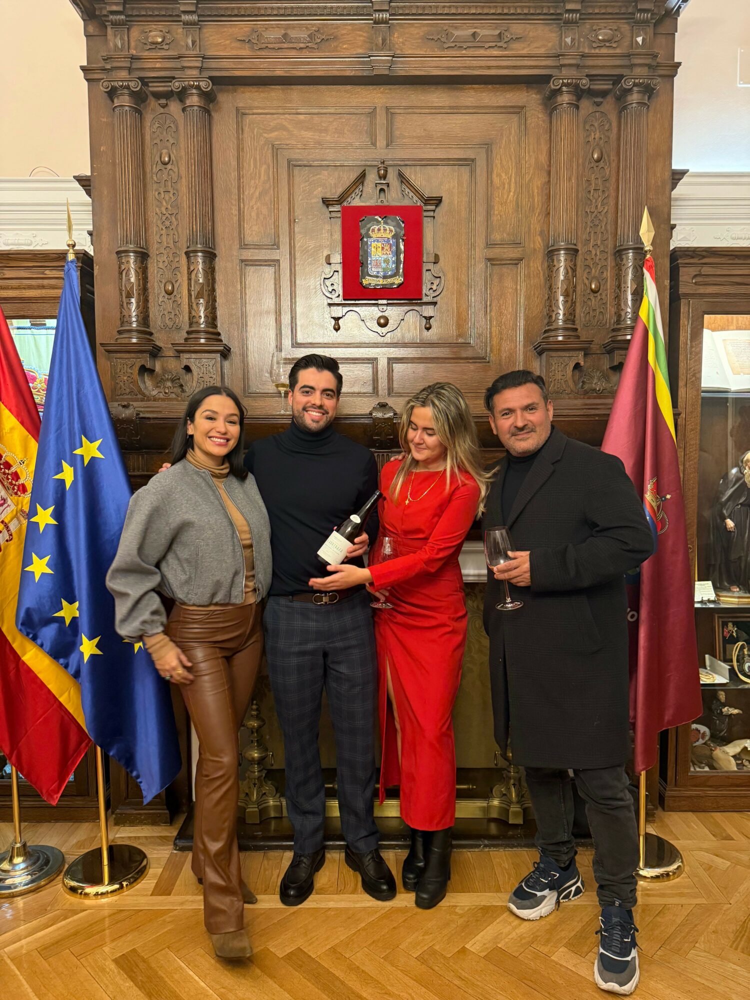 Four people standing in front of a wooden cabinet with a European Union flag and a red flag, smiling and posing for a photo.