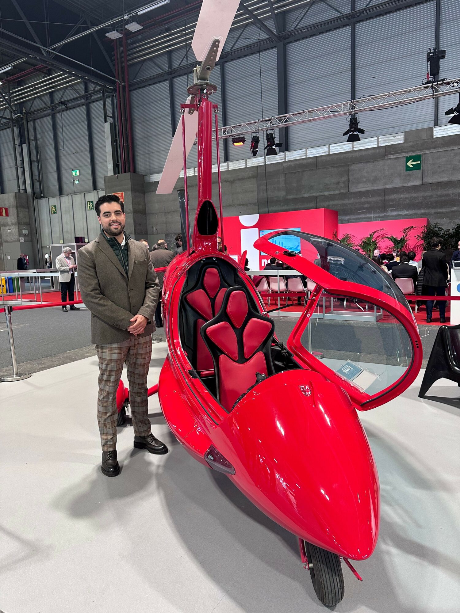 Person standing next to a red, enclosed, three-wheeled vehicle with a transparent canopy at an indoor exhibition.