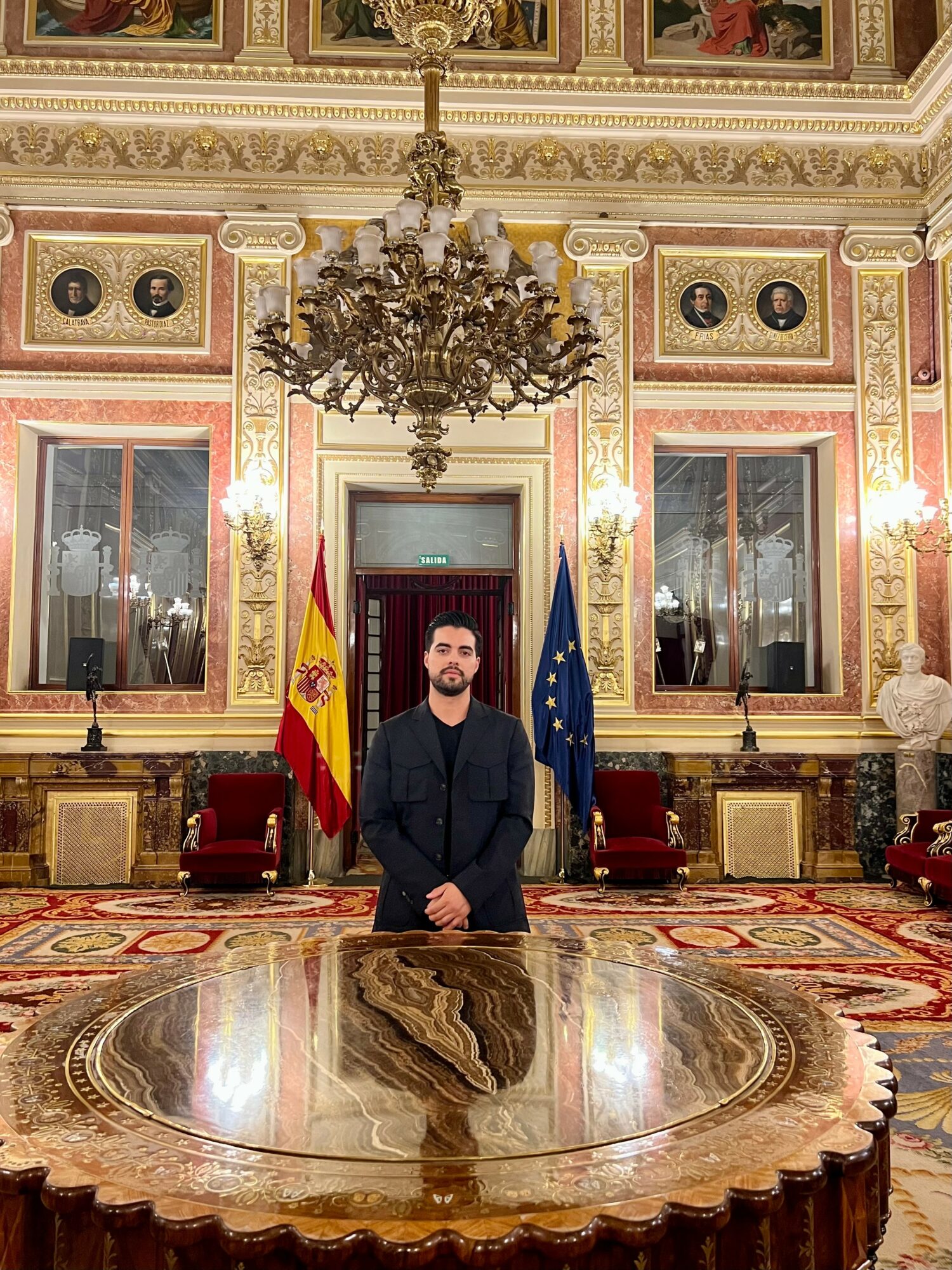 Man standing in a grand ornate room with flags, chandelier, and decorative walls, facing forward with hands clasped.