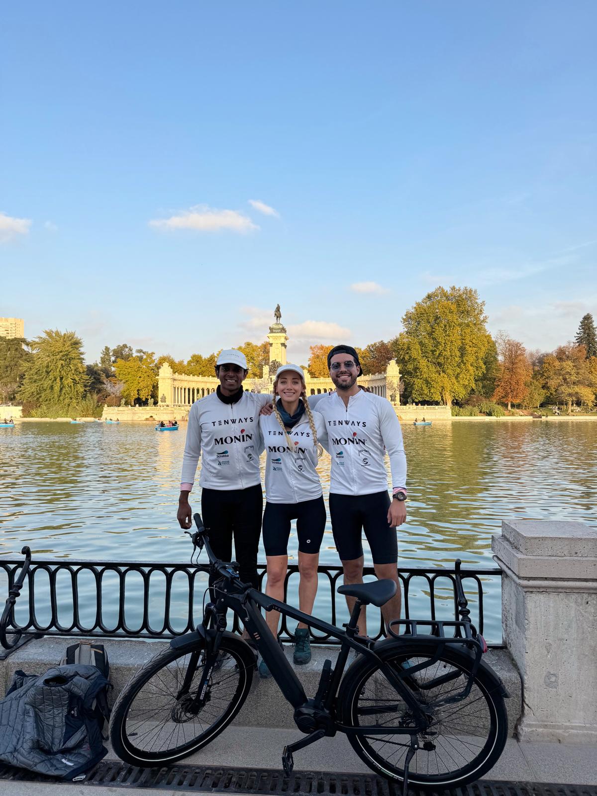 Three people stand by a river with a historic building and trees in the background, smiling and wearing matching shirts.