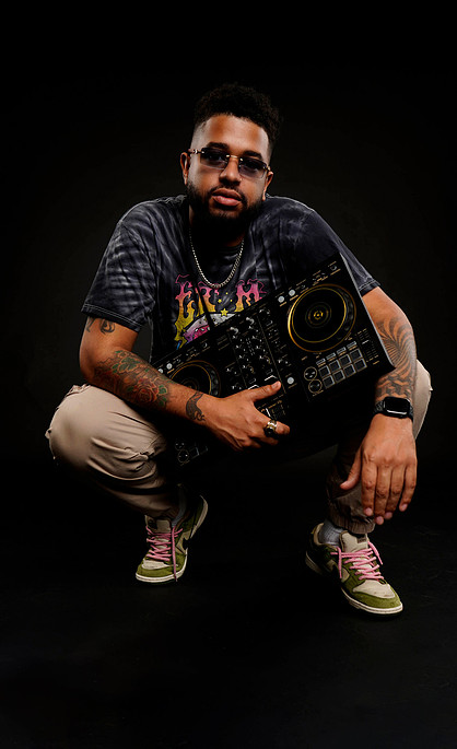 Person crouching with a DJ controller, wearing sunglasses, a dark T-shirt, and beige pants, against a black background.
