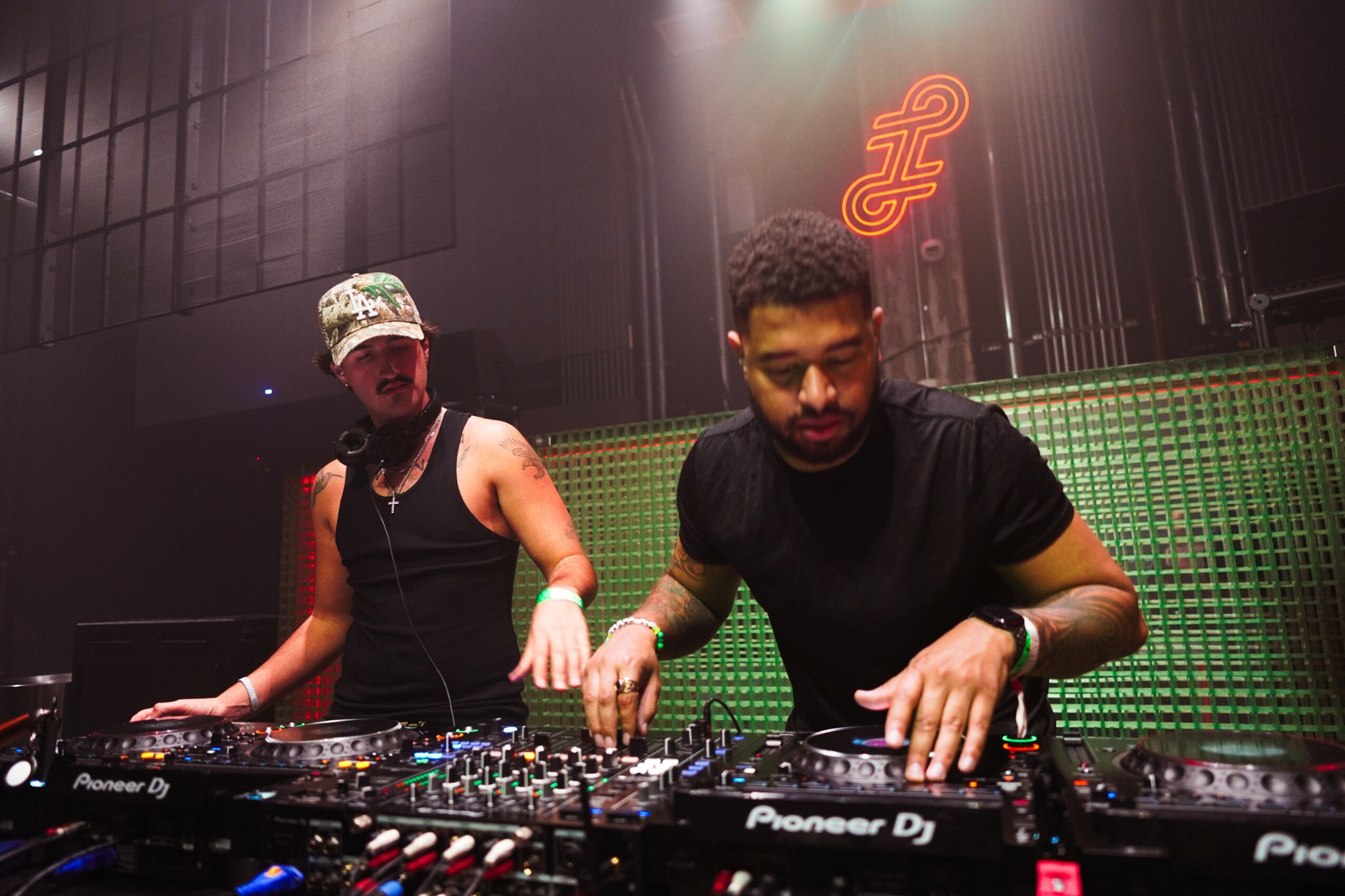 Two DJs perform at a club with green lighting and a neon sign in the background, one wearing a cap and the other with curly hair.