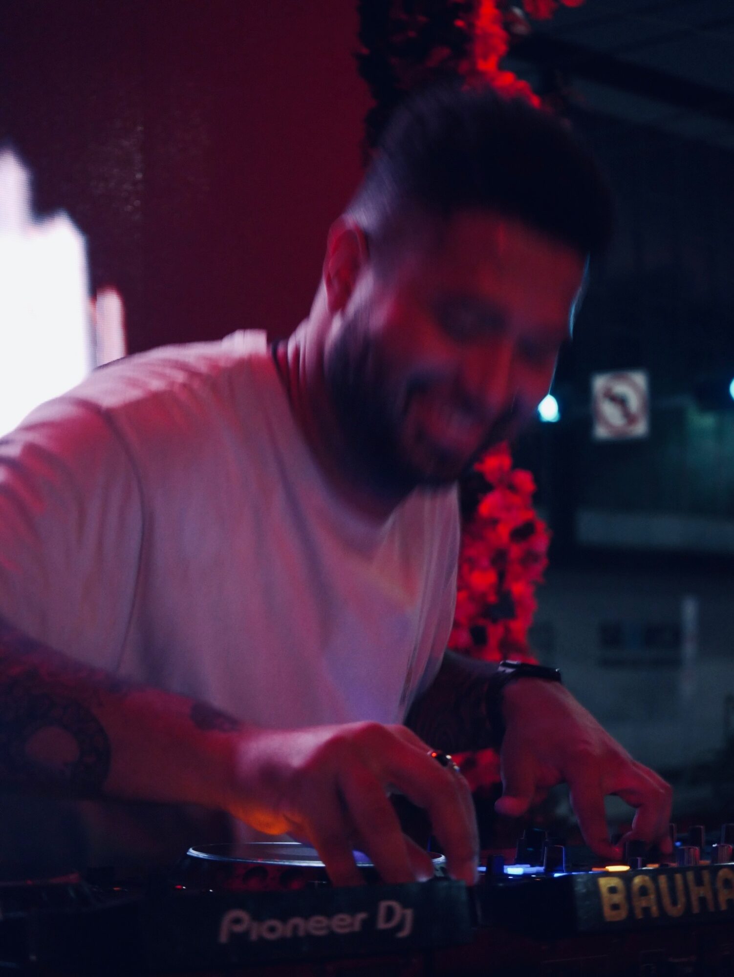 Person with a beard and short hair, wearing a light-colored shirt, appears to be DJing in a dimly lit environment.