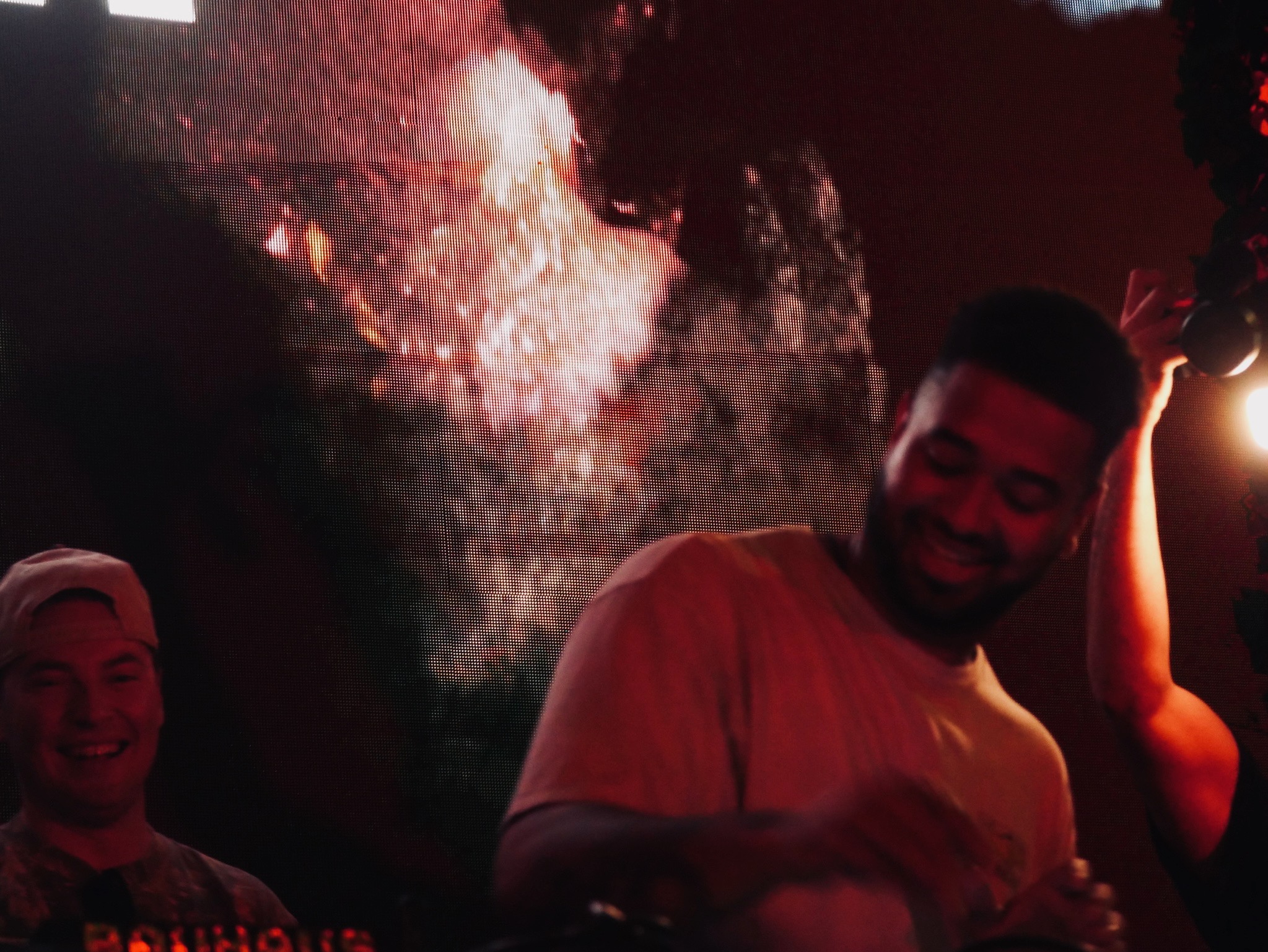 Two people smiling in a dimly lit environment with a large screen or display behind them.